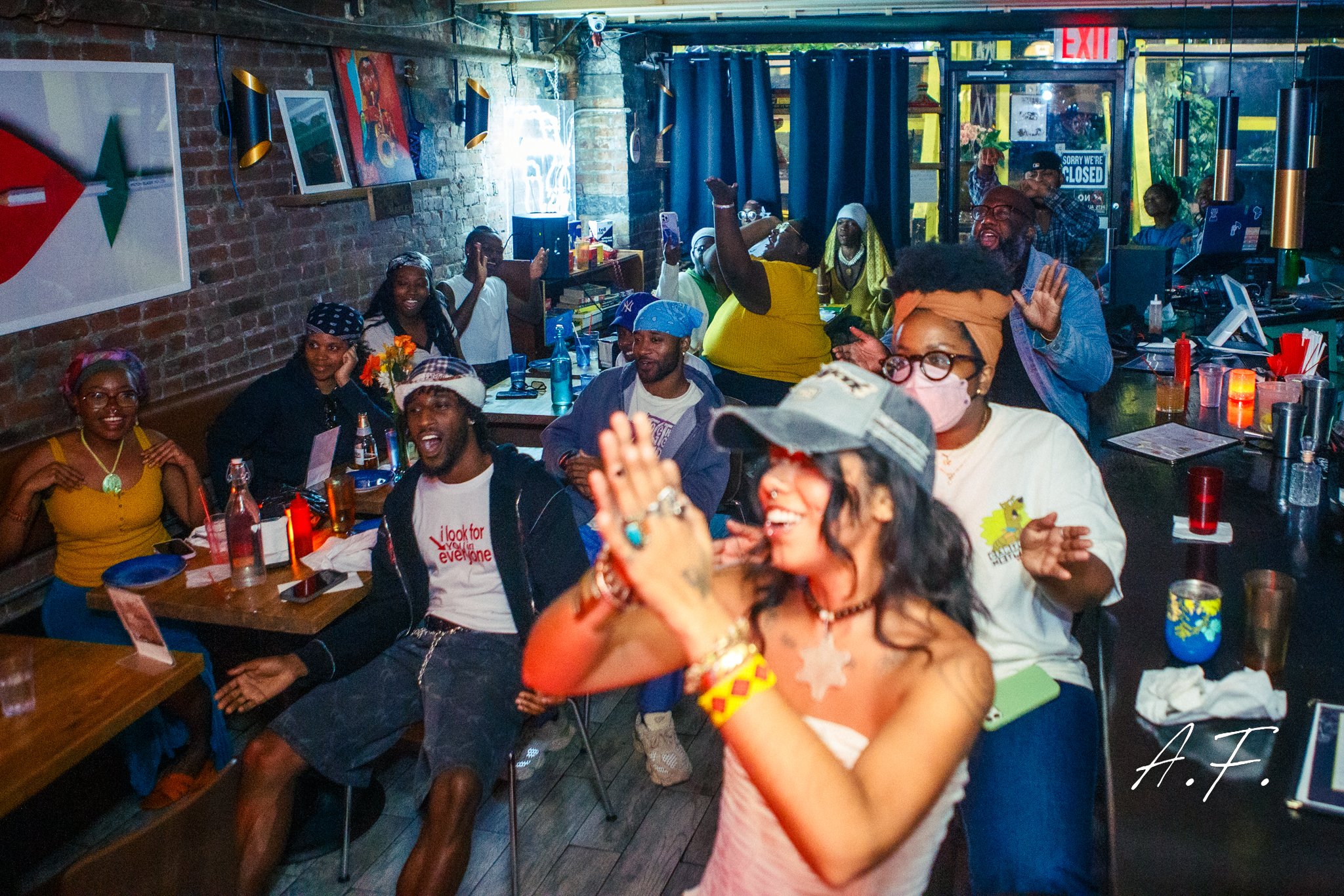 A lively indoor gathering with diverse people enjoying a celebration, sitting at tables with drinks and decorations, some smiling and raising their hands, with a brick wall, artwork, and a window in the background.