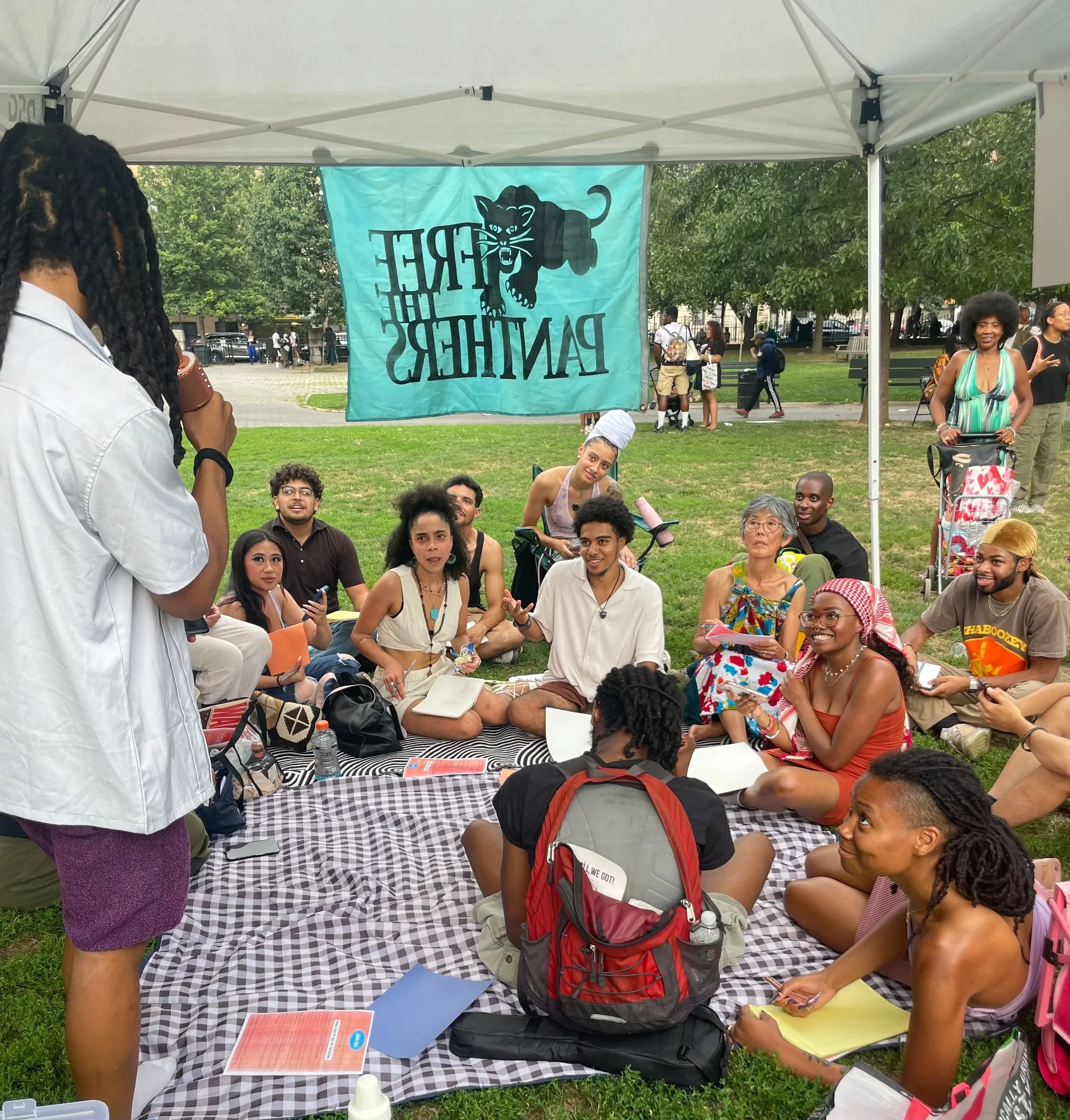 A diverse group of people sitting on a checkered blanket under a tent with a Panther Coffee banner, participating in a discussion in a park.