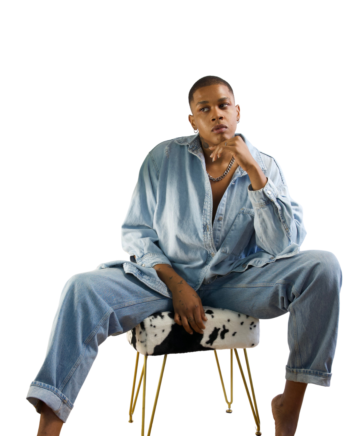 A person sitting on a cow-print chair, wearing an oversized light blue denim jacket and matching jeans, with short hair, earrings, a chain necklace, and tattoos visible on their left arm, with a thoughtful expression.