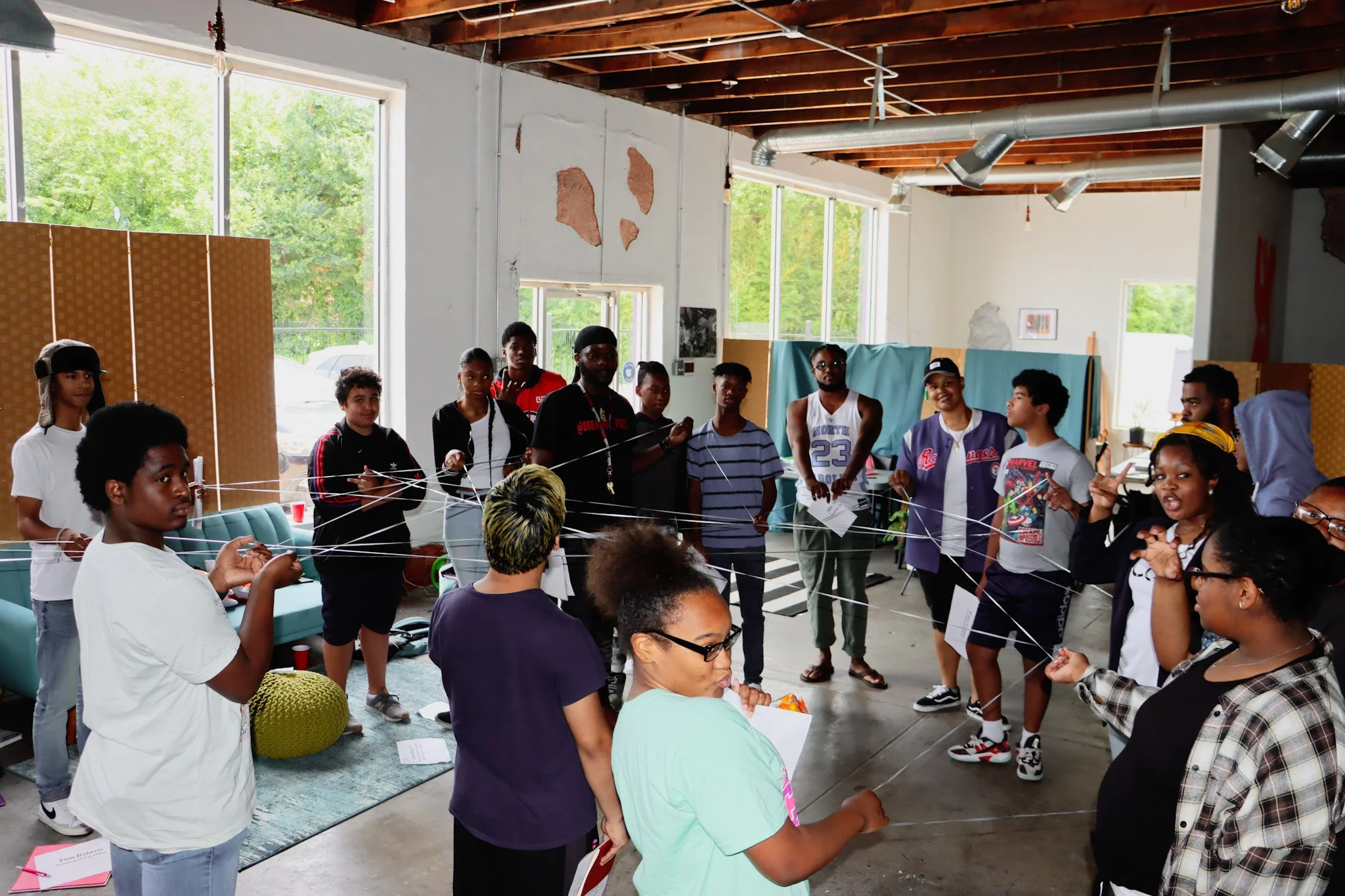 Group of people standing in a circle, holding strings in a team-building activity inside a bright, industrial-style room with large windows and exposed ceiling beams.