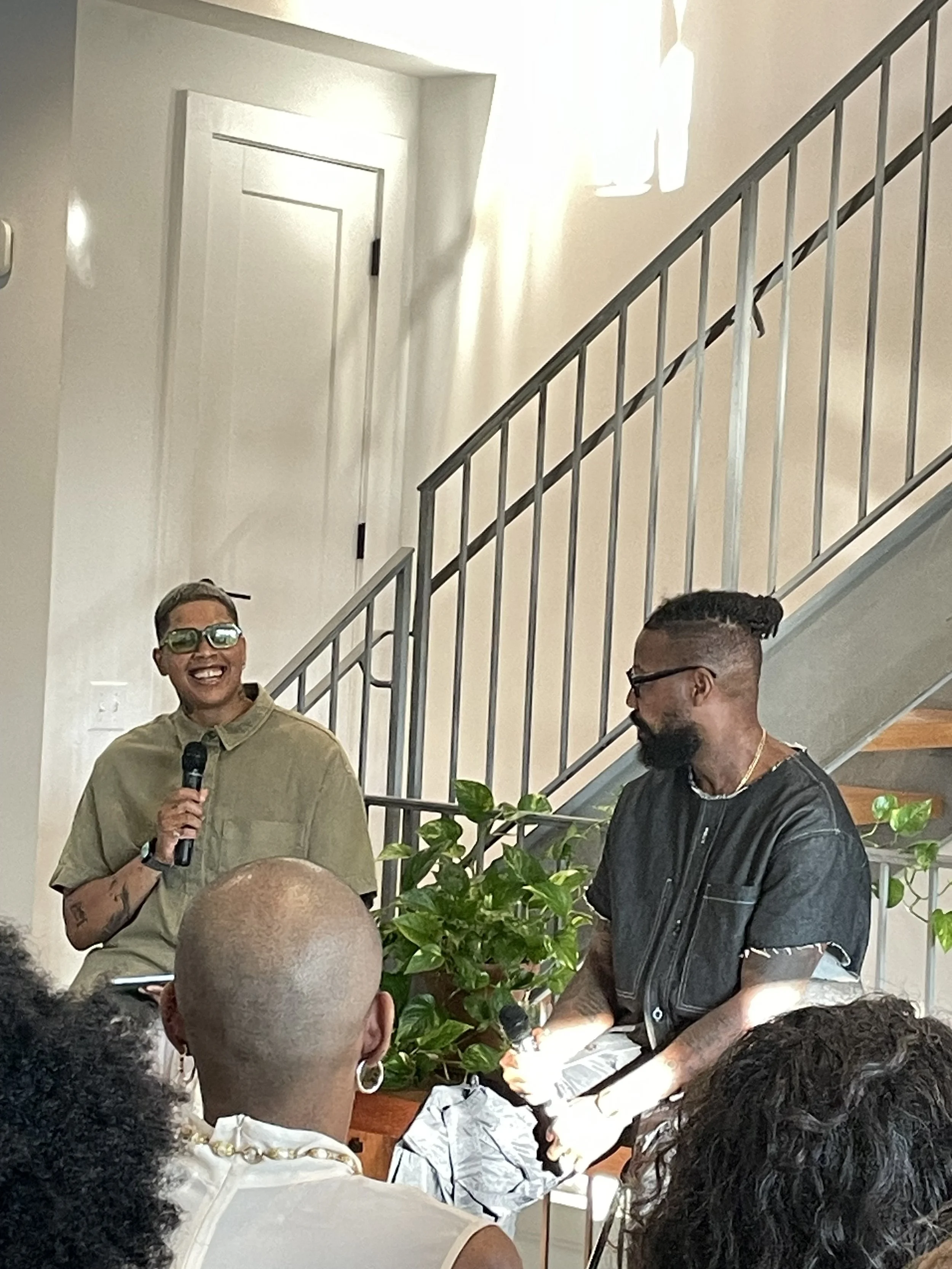 Two men are speaking in front of a small audience indoors. One man is smiling and holding a microphone, wearing glasses and a khaki shirt. The other man has a beard, glasses, and a black leather vest, and is holding a microphone. A woman in the foreground with short hair and hoop earrings is listening. There is a green plant between the speakers and a staircase behind them.