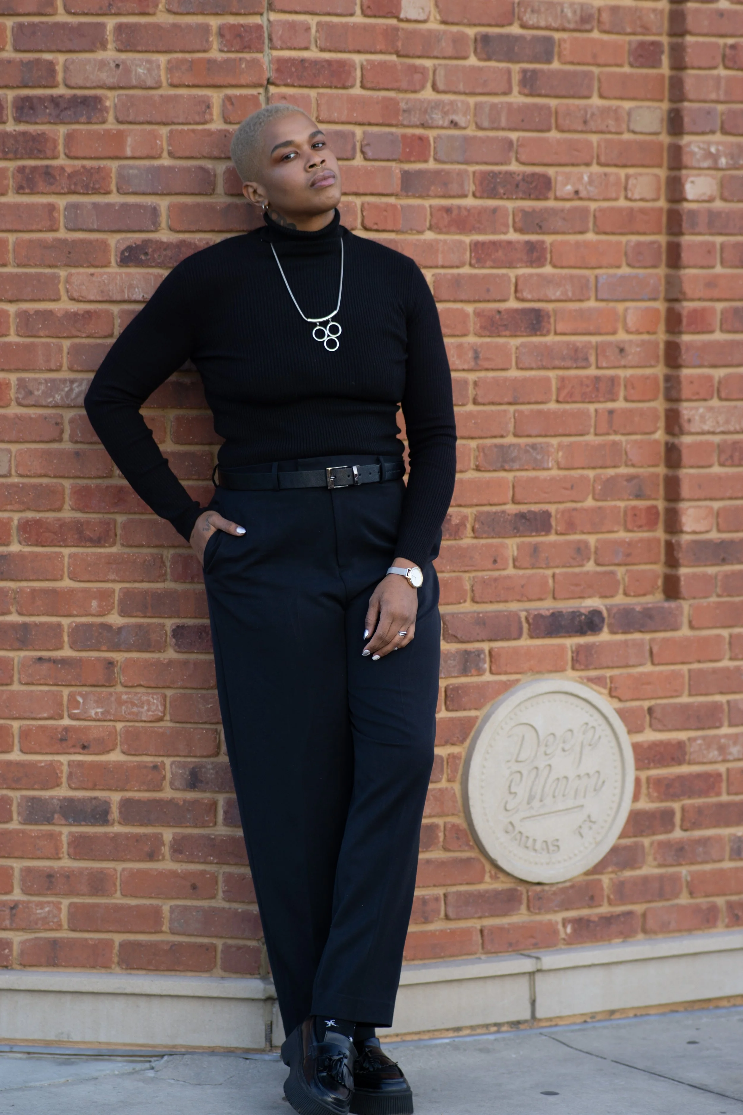 A person with short, platinum blonde hair and medium-dark skin tone standing against a brick wall, wearing a black turtleneck, black high-waisted trousers, black platform shoes, a silver watch, rings, and a necklace.