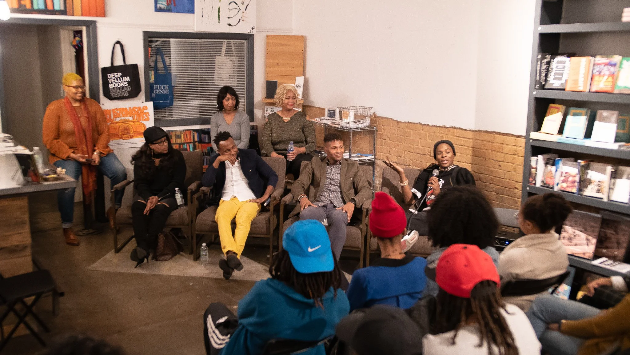 A panel discussion or group event taking place in a bookstore or library setting with seven people seated on chairs and half a dozen audience members in front, some wearing colorful hats.