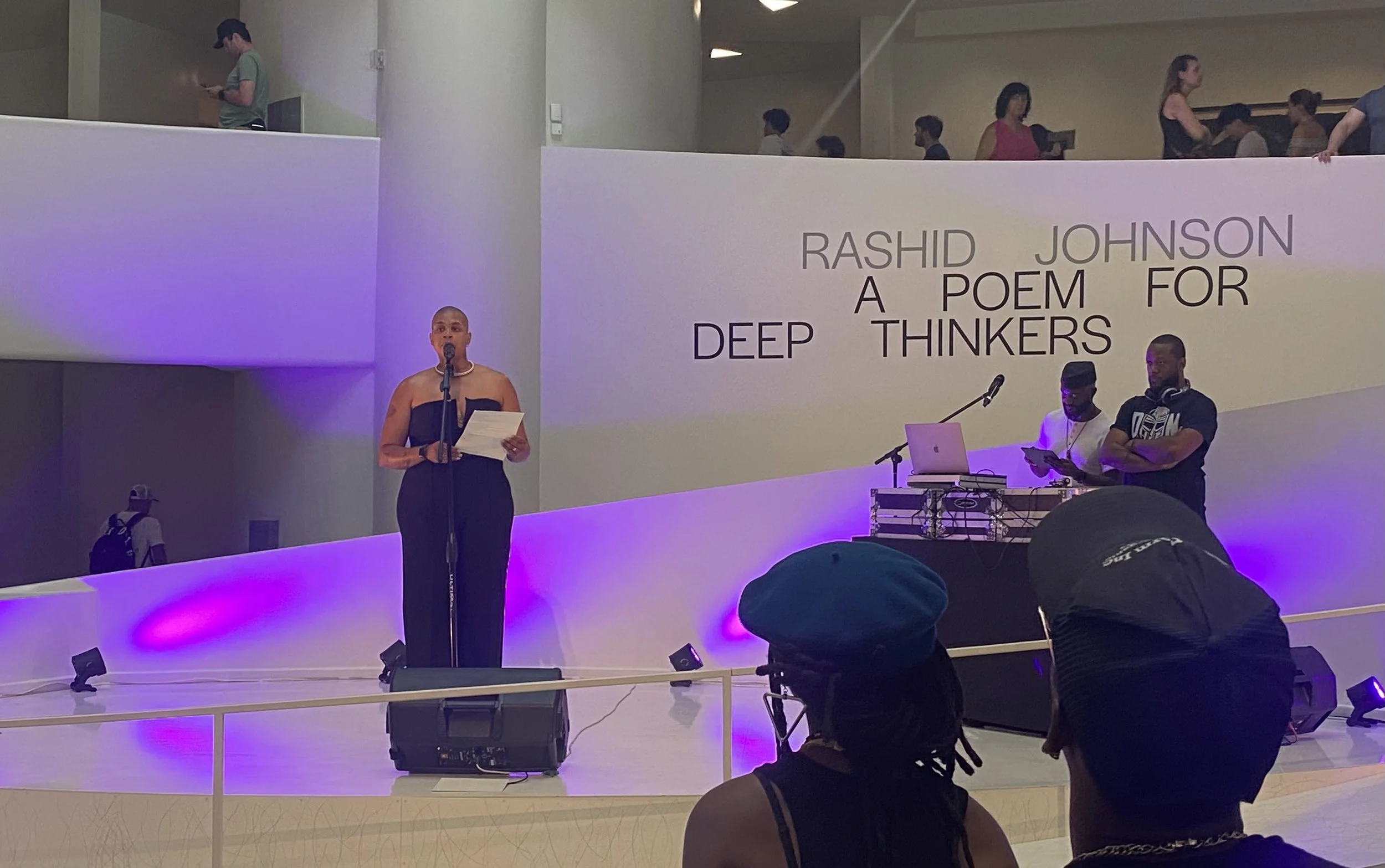A woman is speaking into a microphone on a stage, holding papers. Behind her, a large screen displays the text: 'Rashid Johnson: A Poem for Deep Thinkers.' Two men are standing behind a DJ setup on the right side of the stage, one is using a phone, the other has headphones around his neck. There are audience members watching, some on a balcony level above the stage.