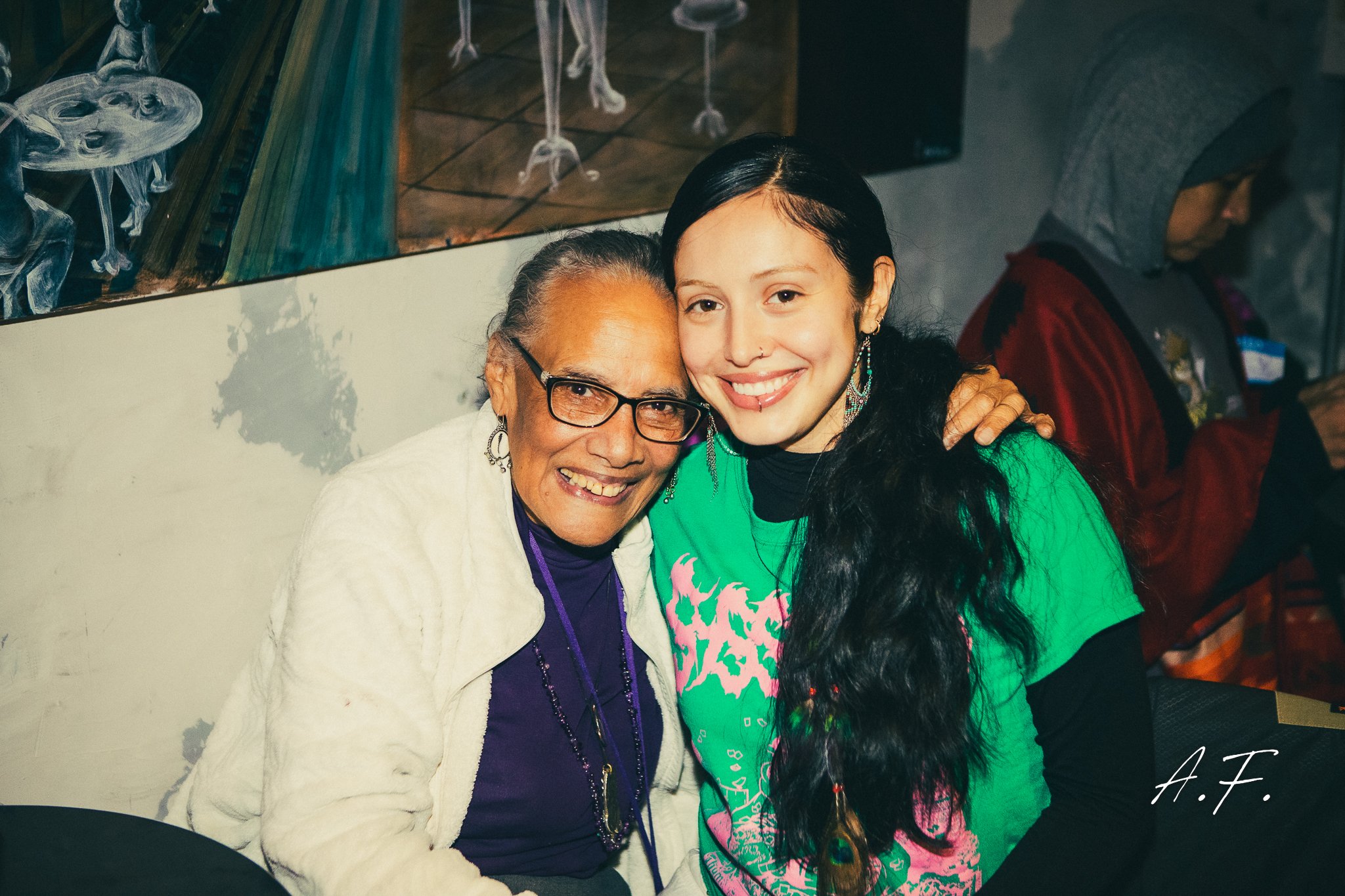 Two women smiling and hugging in an indoor setting. The elderly woman on the left wears glasses, a white jacket, and purple clothing underneath. The younger woman on the right has long black hair, a green t-shirt with pink graphics, and large earrings.