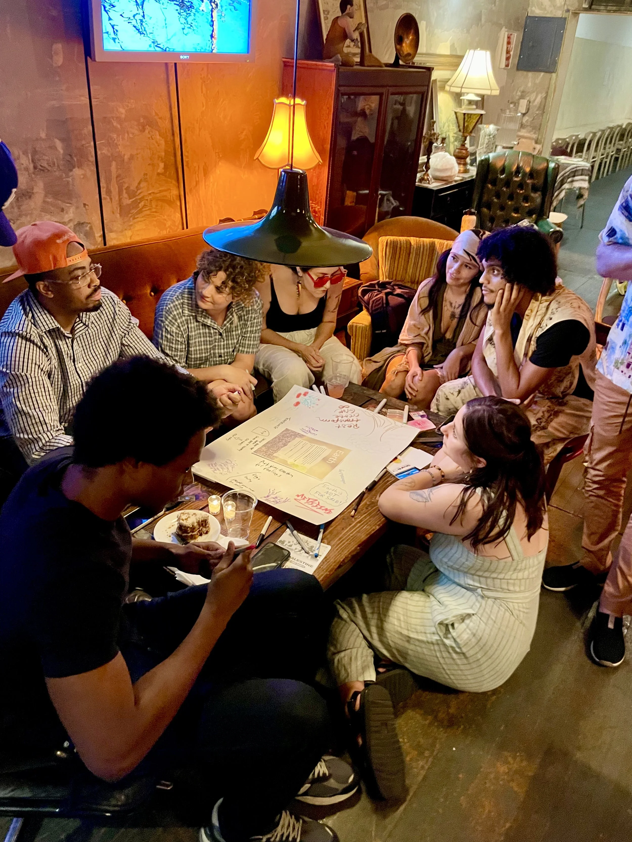 Group of people gathered around a table with colorful papers, drawings, and markers in a cozy, dimly lit room, possibly hosting a social or creative event.