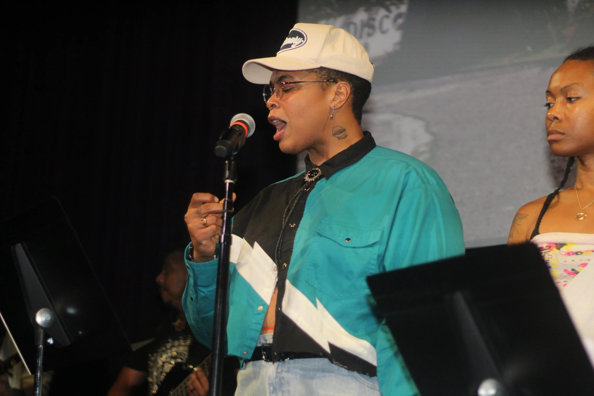 Woman with short hair, glasses, and tattoos singing into a microphone on stage, wearing a colorful jacket and a beige cap, with another woman in the background.