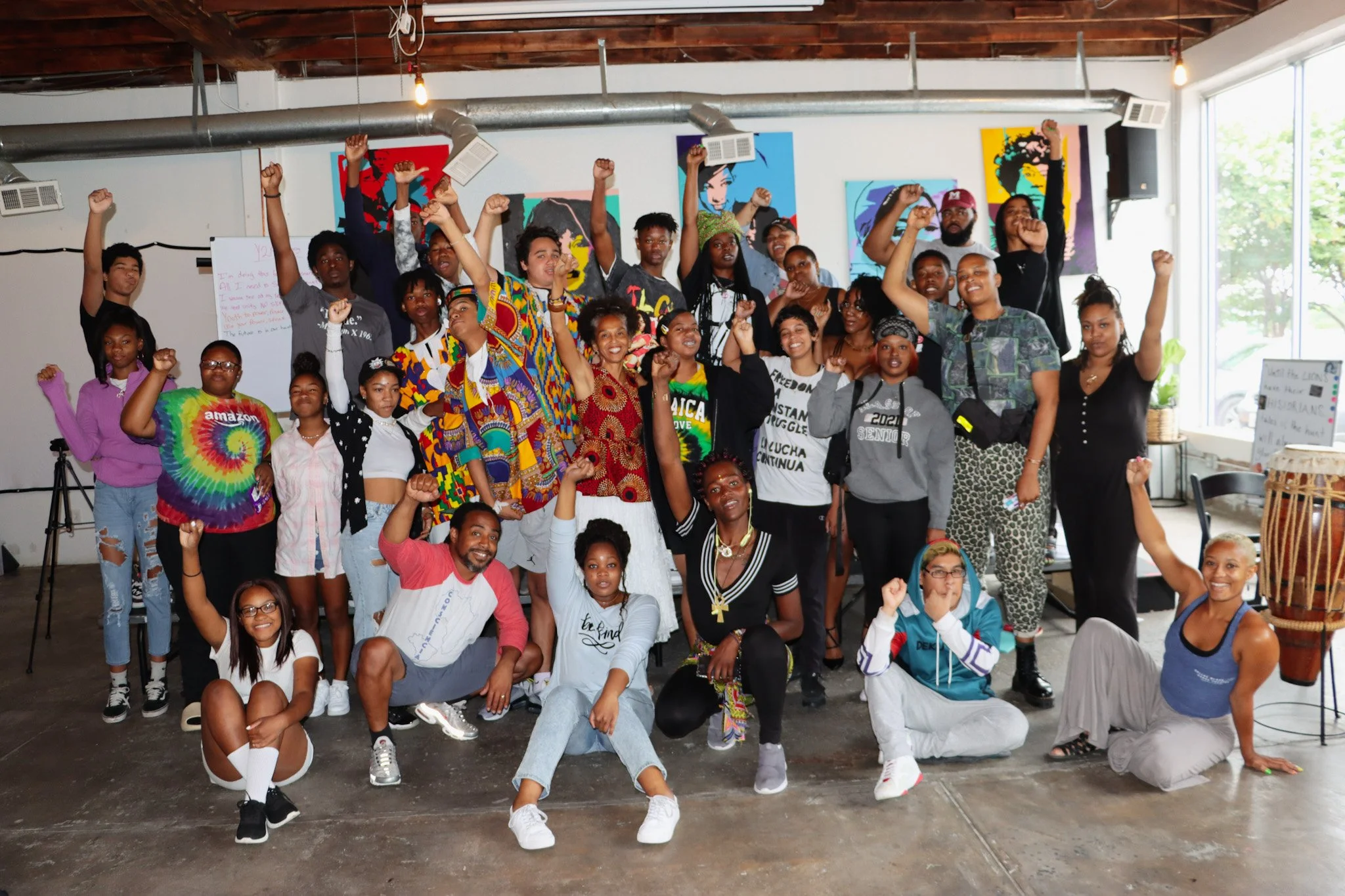A large diverse group of people, including children, teenagers, and adults, gathered indoors in a spacious, well-lit room with art on the walls, raising their fists in celebration or solidarity.