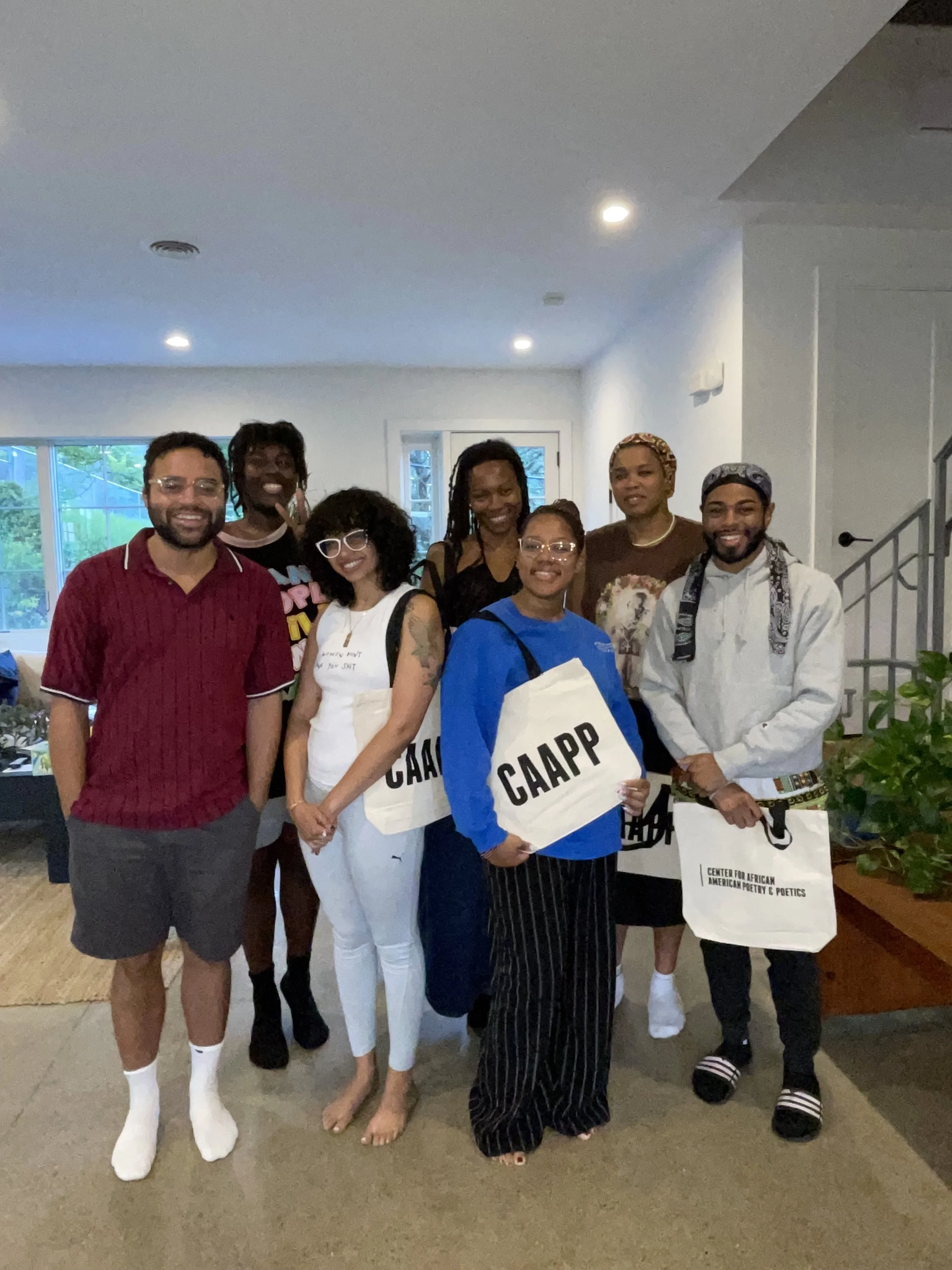 A group of nine diverse people standing together indoors, smiling, some holding tote bags with 'CAAPP' and 'Center for African American Poetry & Politics' printed on them.