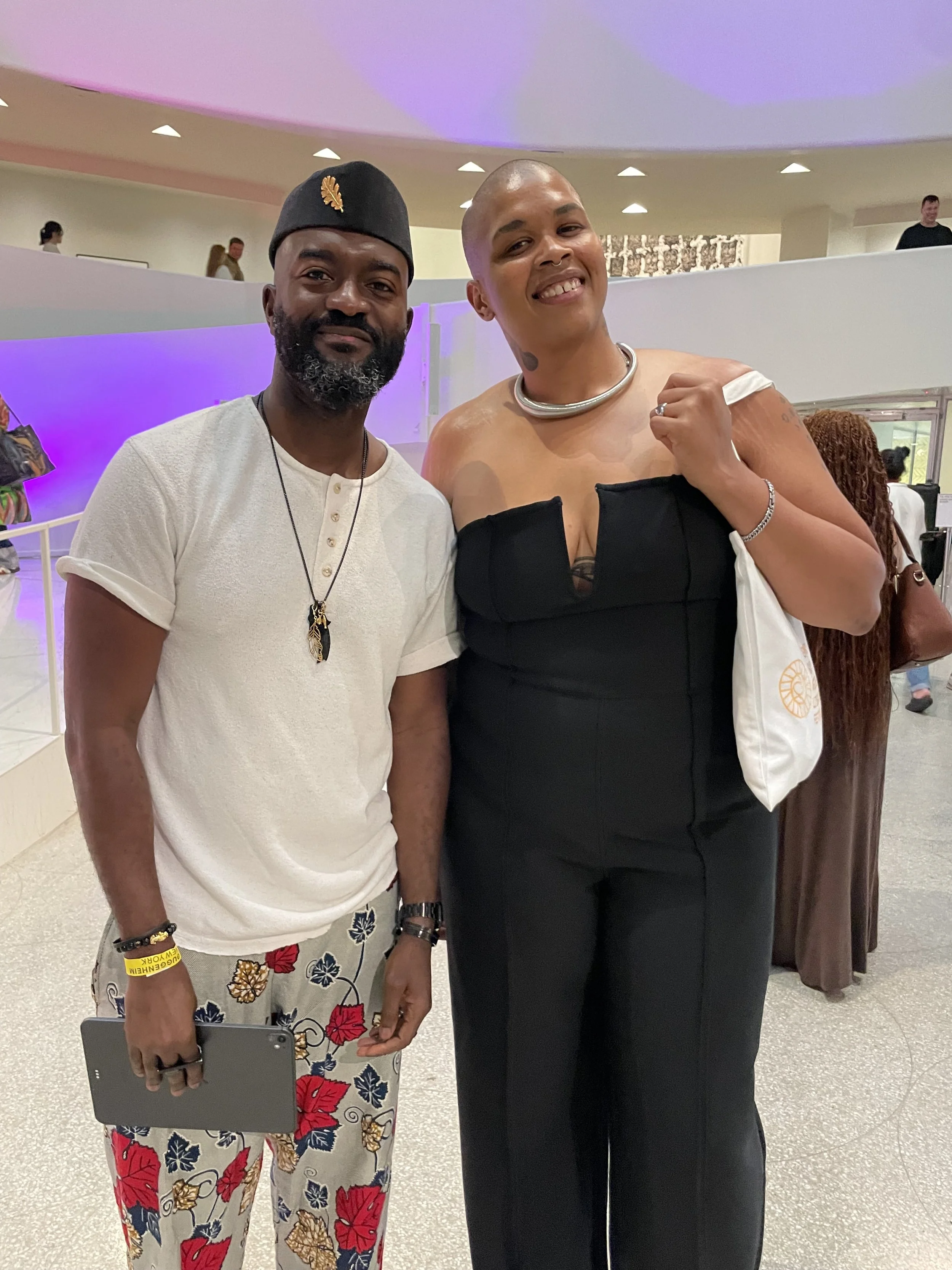 A man wearing a black cap, white shirt, and floral pants standing next to a woman in a black dress, both smiling at the camera inside a brightly lit venue with curved walls and purple lighting.