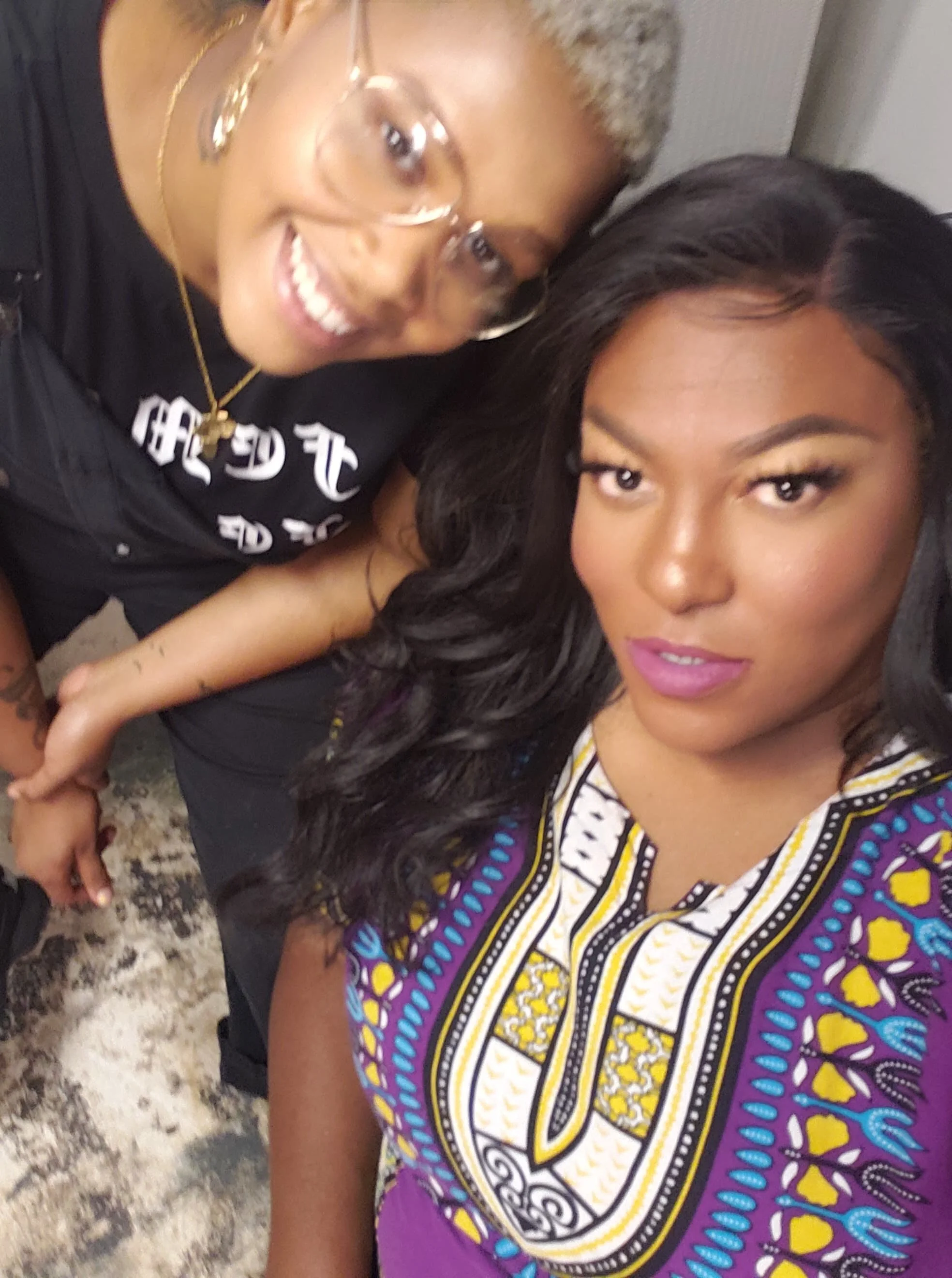 Two women taking a close-up selfie, one with long black hair and purple lipstick, and the other with short blonde hair and glasses, both smiling.