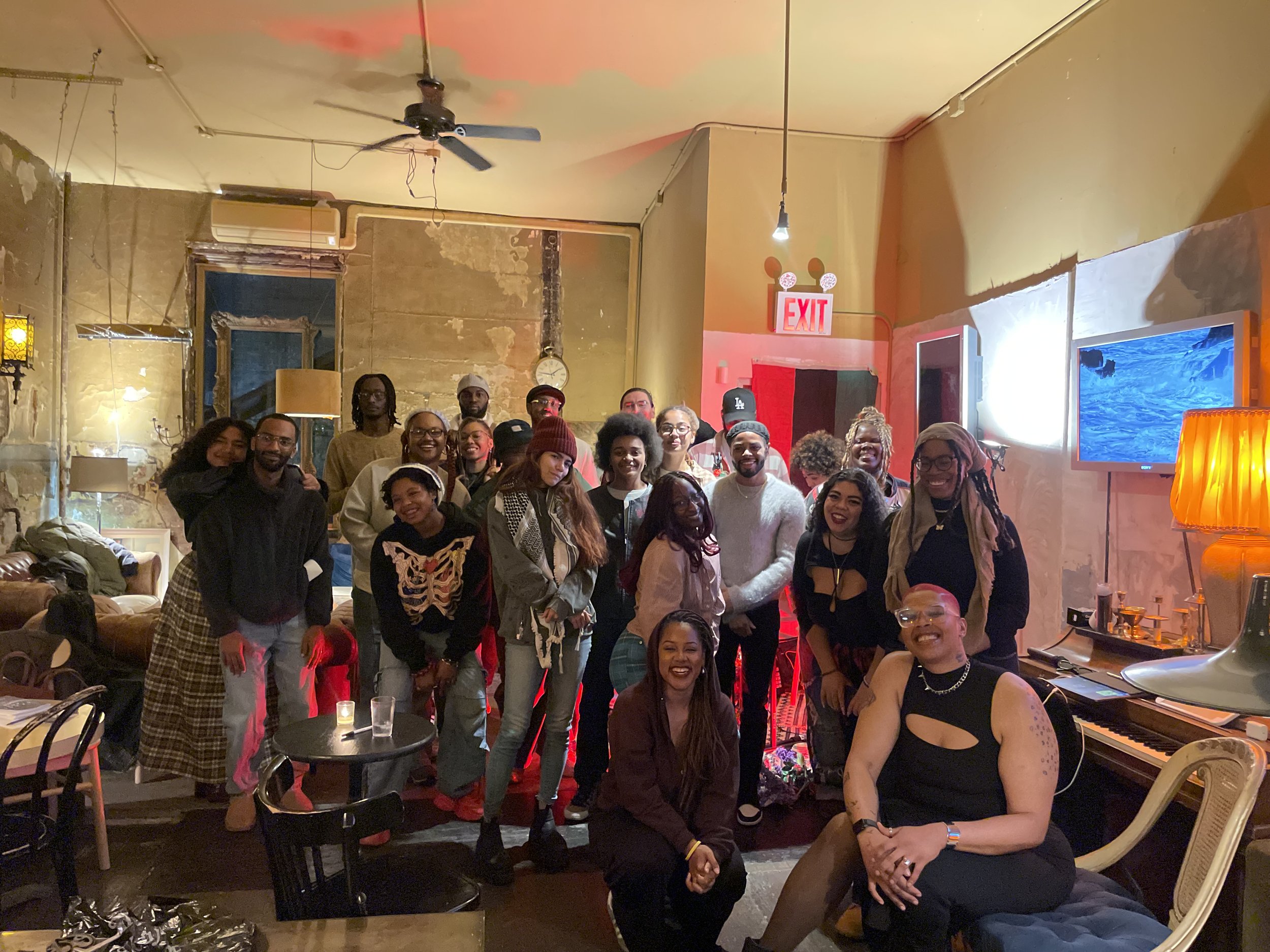 Group of people gathered indoors in a cozy, eclectic space with warm lighting, vintage decor, and a mix of casual and expressive clothing. Some are smiling and posing for the photo.