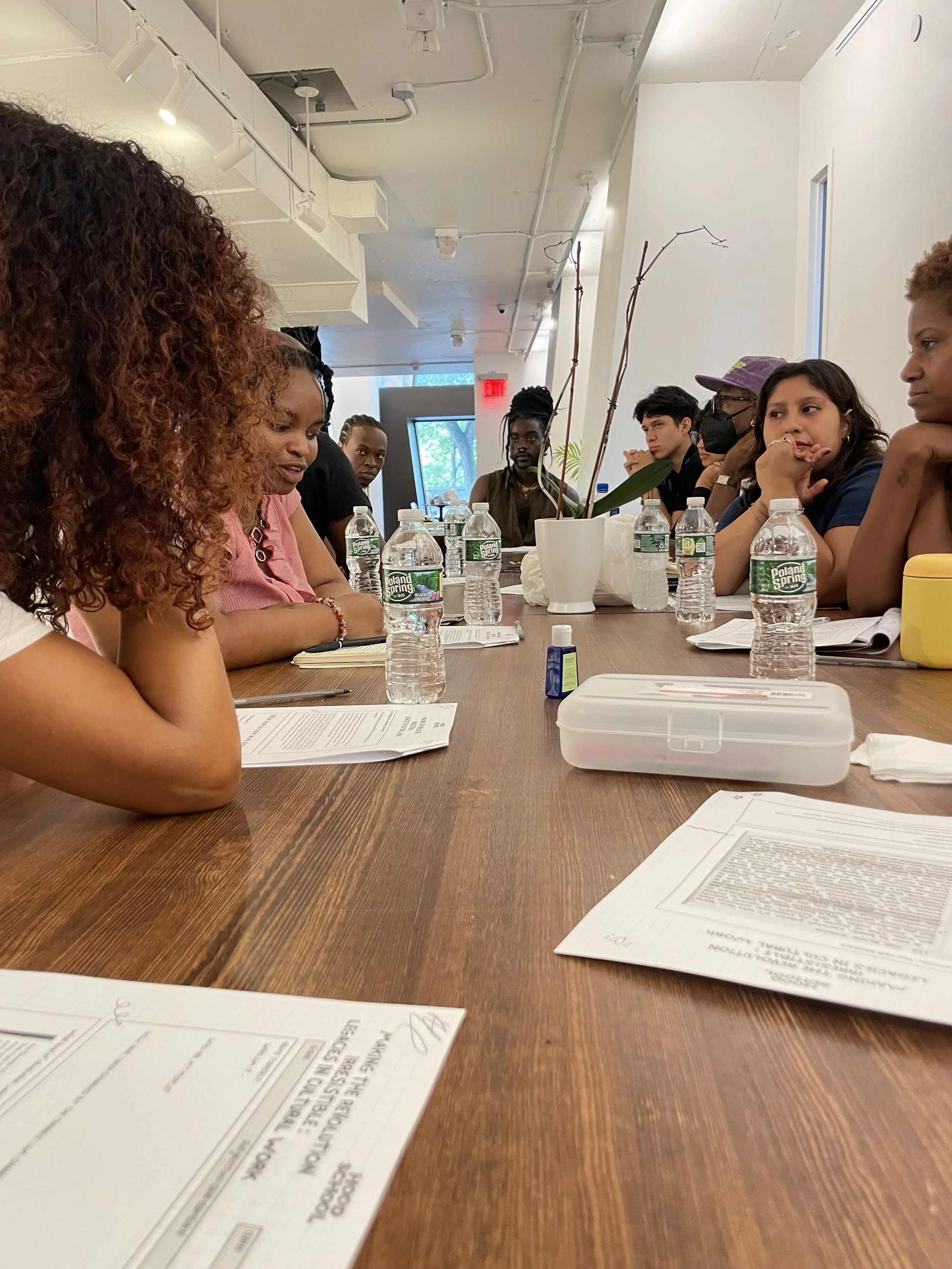 A diverse group of people sitting around a long wooden table during a meeting or discussion in a bright, modern room with white walls and ceiling. There are water bottles, papers, and a potted plant on the table.