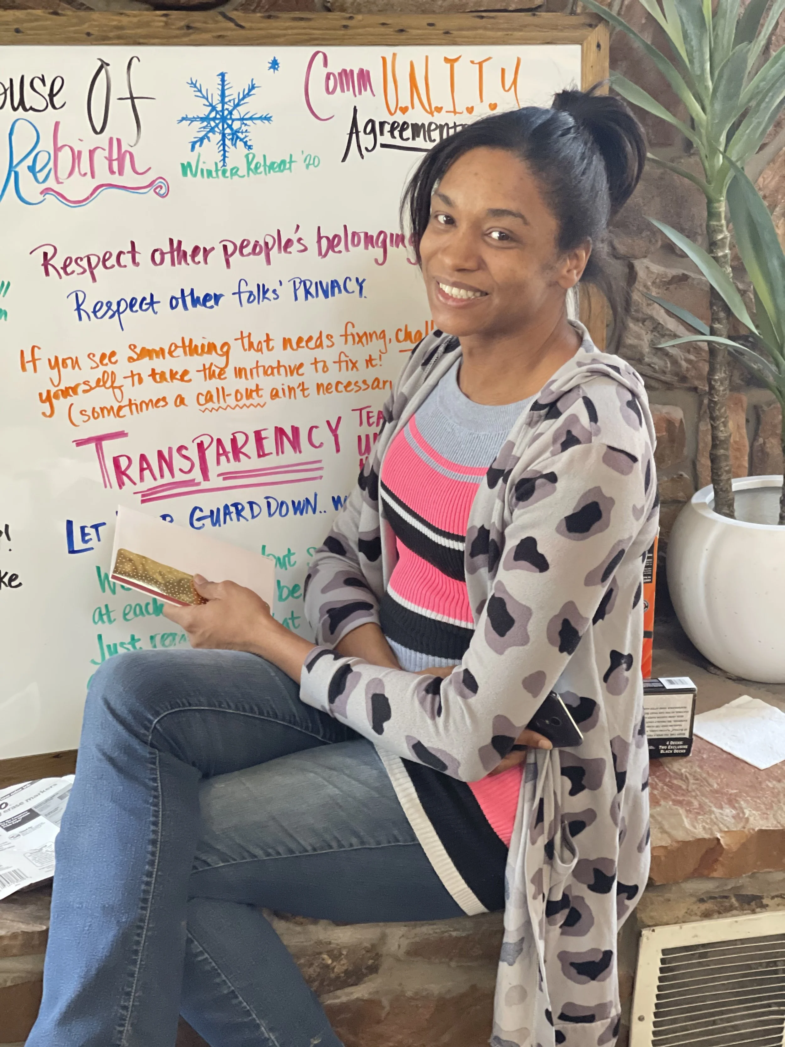 A woman sitting on a stone ledge in front of a colorful whiteboard with writing about community agreements. She has dark hair tied back, is smiling, and is holding a gold-foil wrapped item in her right hand while her left hand rests on her knee. The background includes a brick wall and a potted plant.