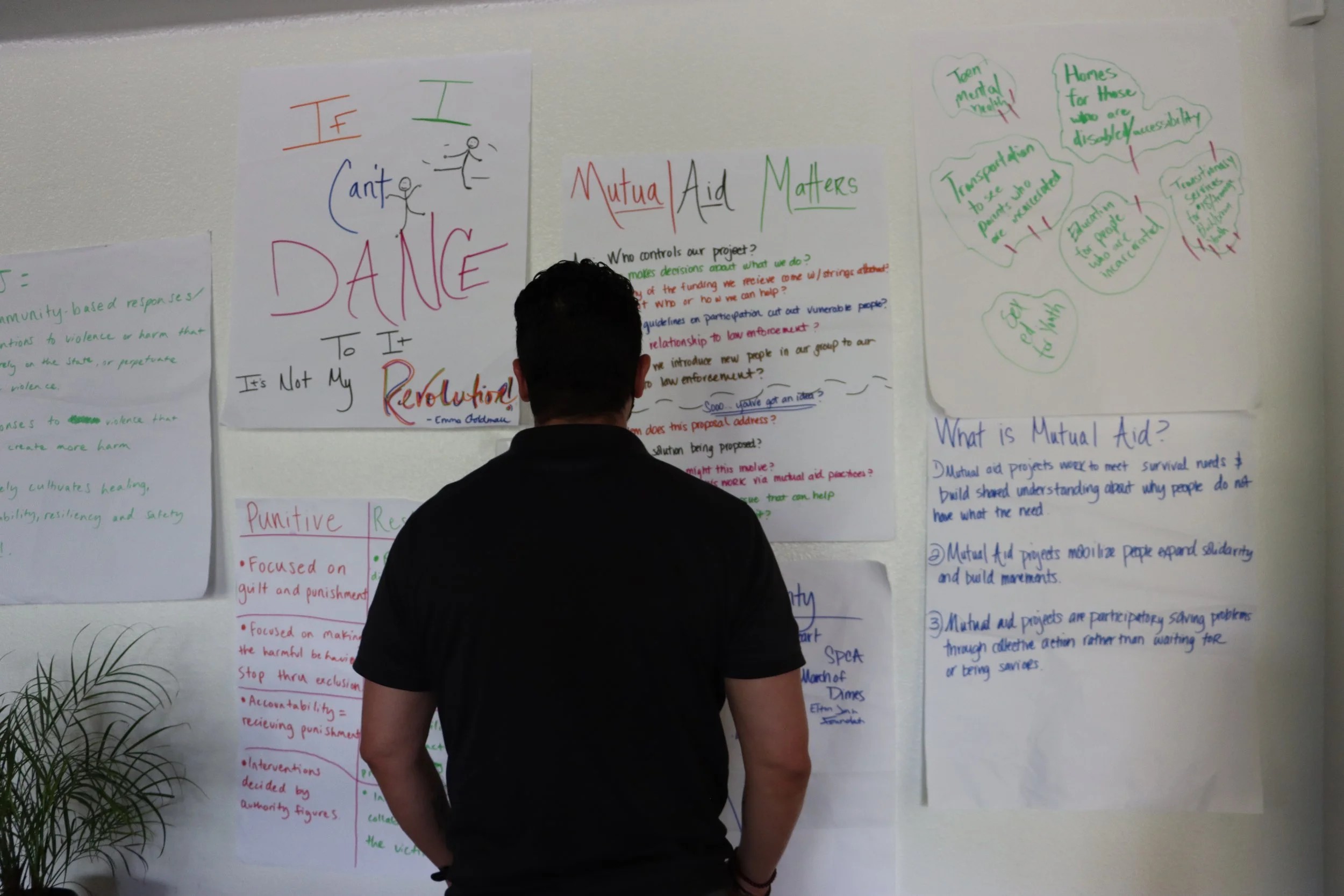 A person standing in front of a wall covered with various colorful posters and handwritten notes about mutual aid and social justice, reading the information displayed.