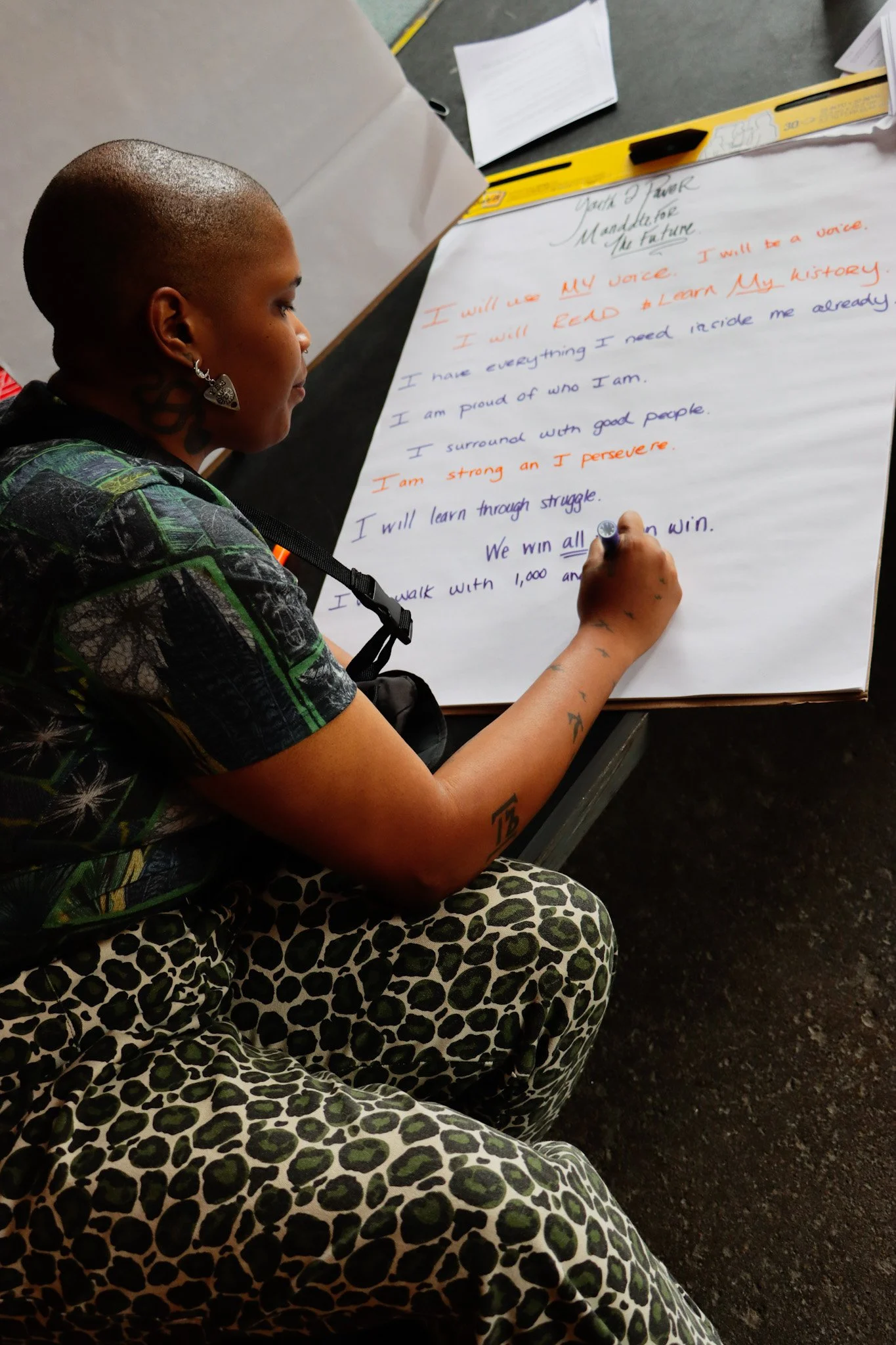 A person sitting at a table and writing on a large sheet of paper with a marker. The person has a shaved head, tattoos on their arm, earrings, and is wearing a patterned shirt and leopard-print pants. The paper contains a motivational speech written in colorful ink.