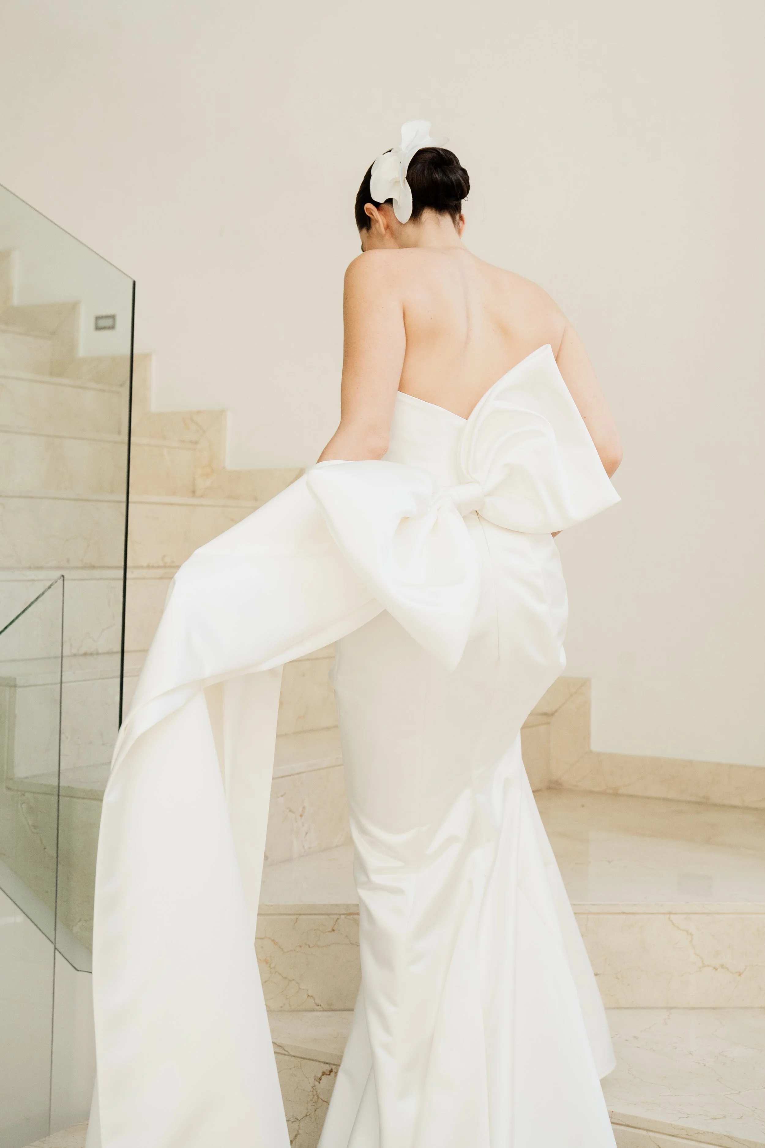 A woman in a white strapless gown with a large bow at the back, standing on marble stairs, with her back to the camera, her hair styled with a white hair accessory.