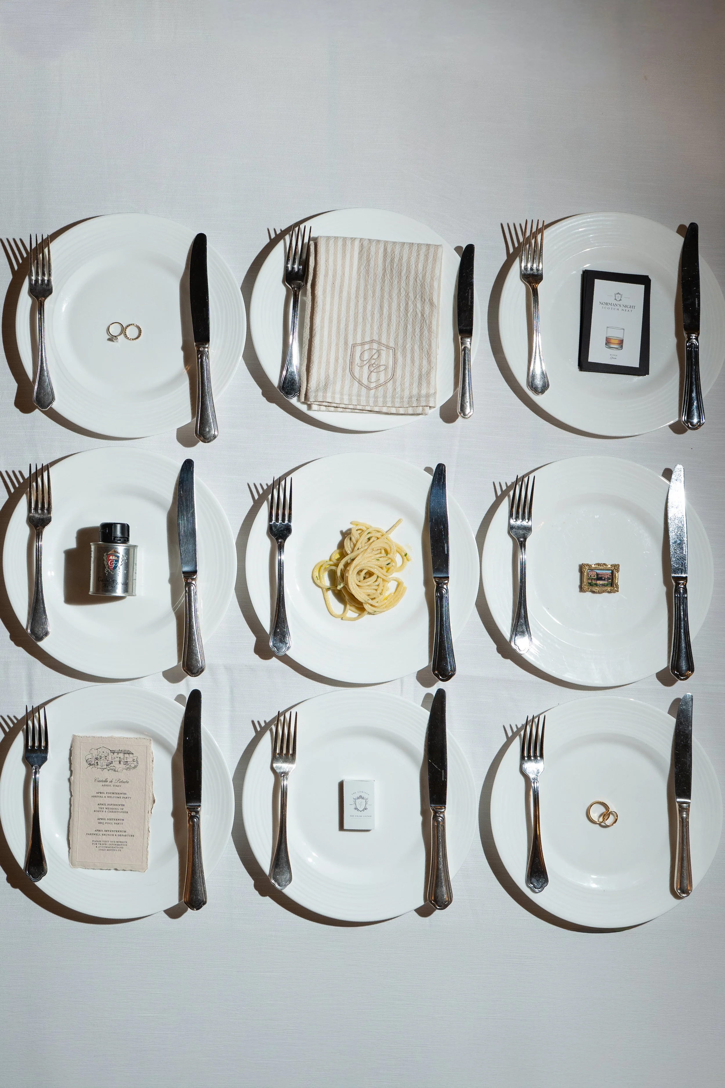 Table setting with white plates, silverware, and various small items on a white tablecloth. Top row, from left to right: two wedding rings, a white plate with a knife and fork, a napkin with embroidery, and a menu card. Bottom row, from left to right: a silver lighter, a plate of pasta, a small framed picture, and two wedding rings.