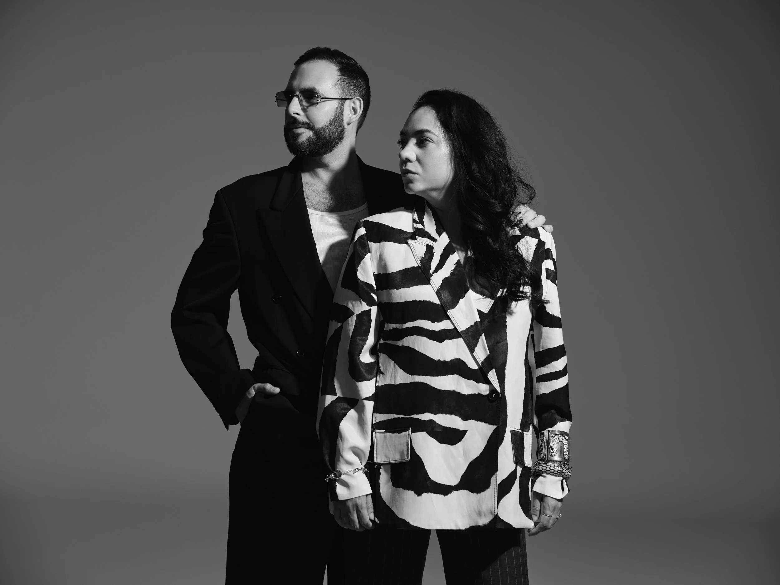 Black and white photo of a man and woman standing close, with the man’s arm around the woman’s shoulder, both looking away from the camera against a plain background.