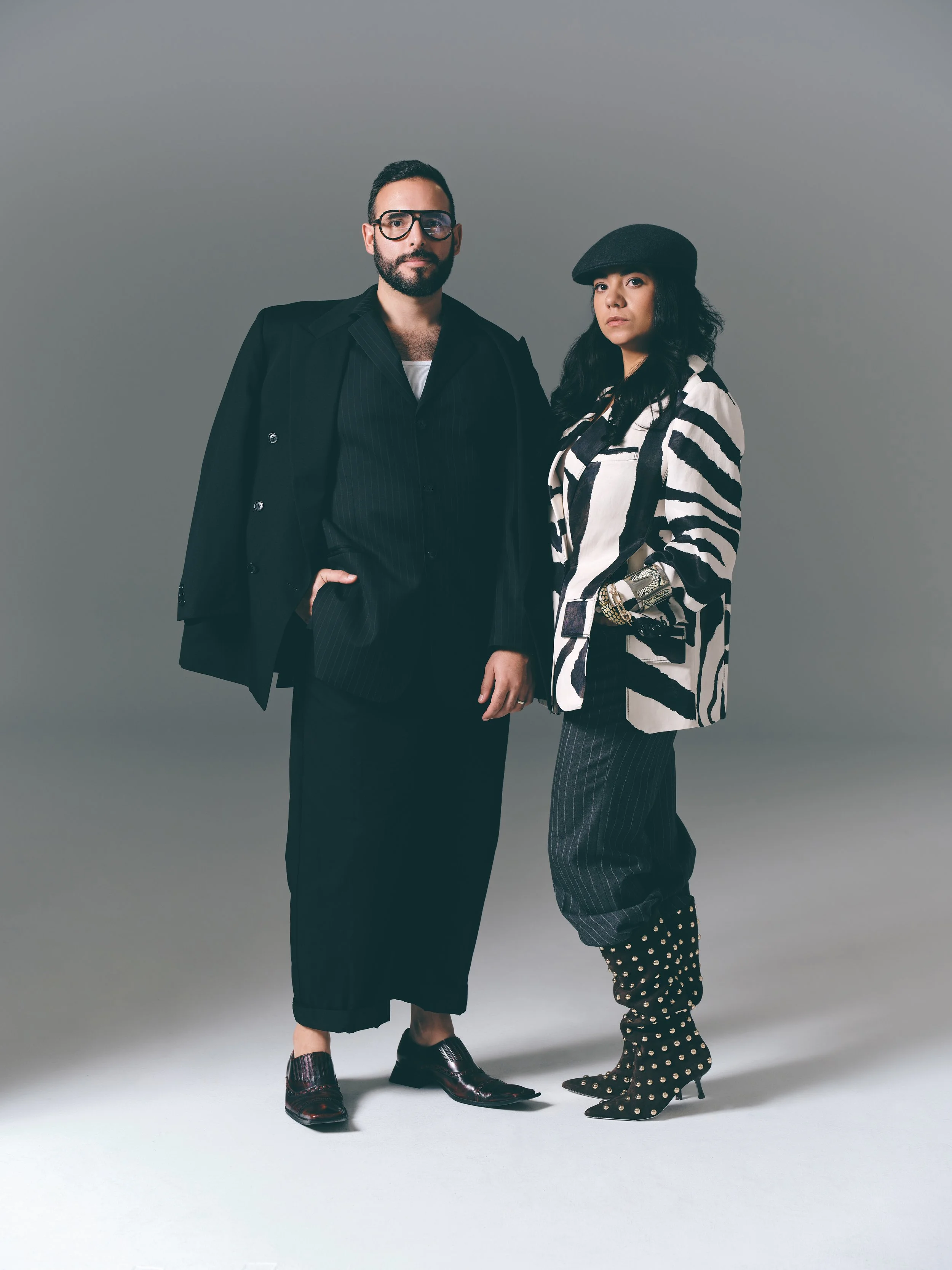 Two stylish individuals standing against a plain gray backdrop. The man on the left has dark hair, a beard, and wears glasses, a black pinstripe suit with an open collar shirt, and black dress shoes. The woman on the right has long black hair, a black beret, a black and white zebra-striped blazer, black pinstripe pants, and black heeled boots with gold studs.