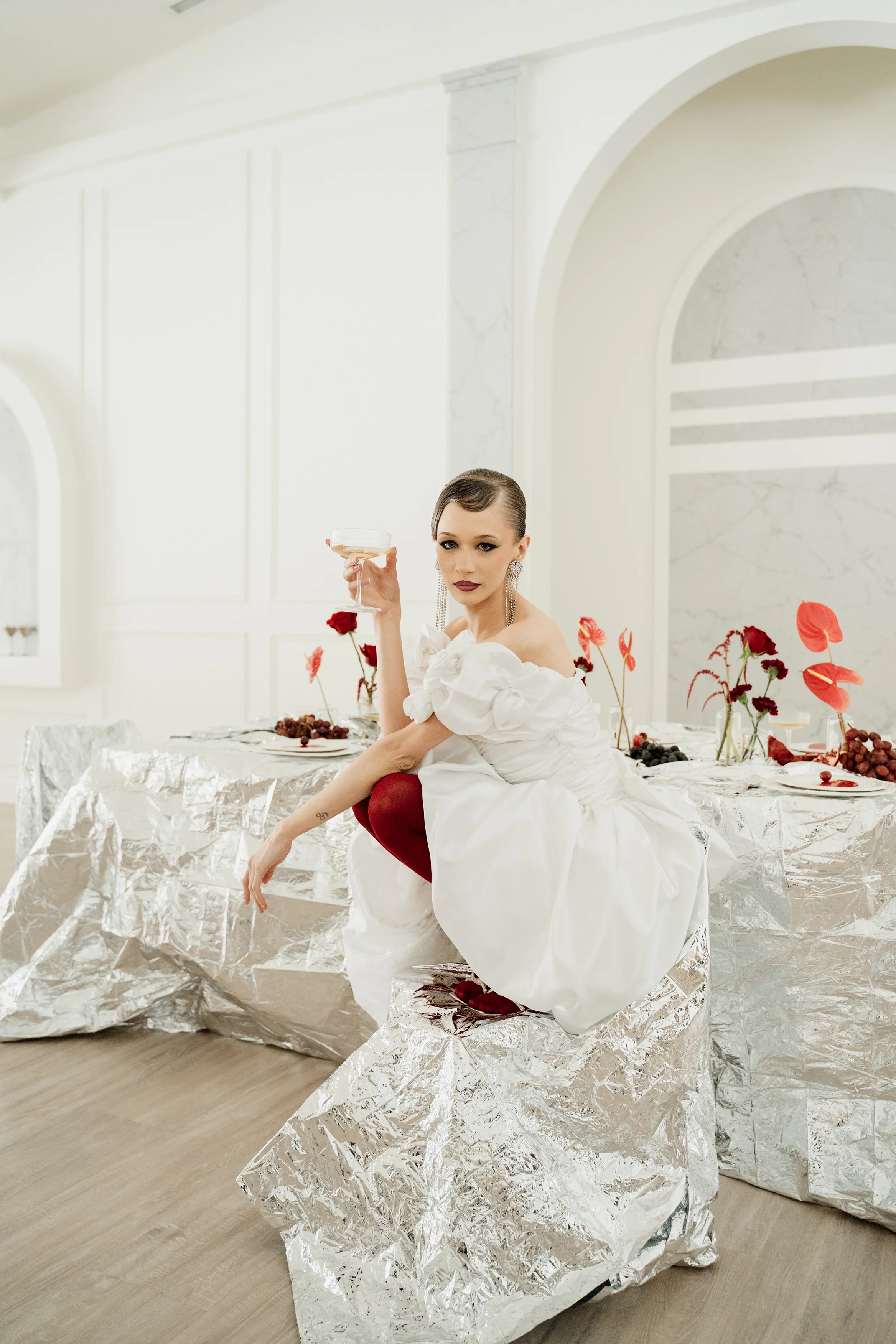 A woman dressed in a white dress sitting on a silver foil-covered chair at a table adorned with red flowers and grapes, holding a glass of champagne in a bright, modern room.