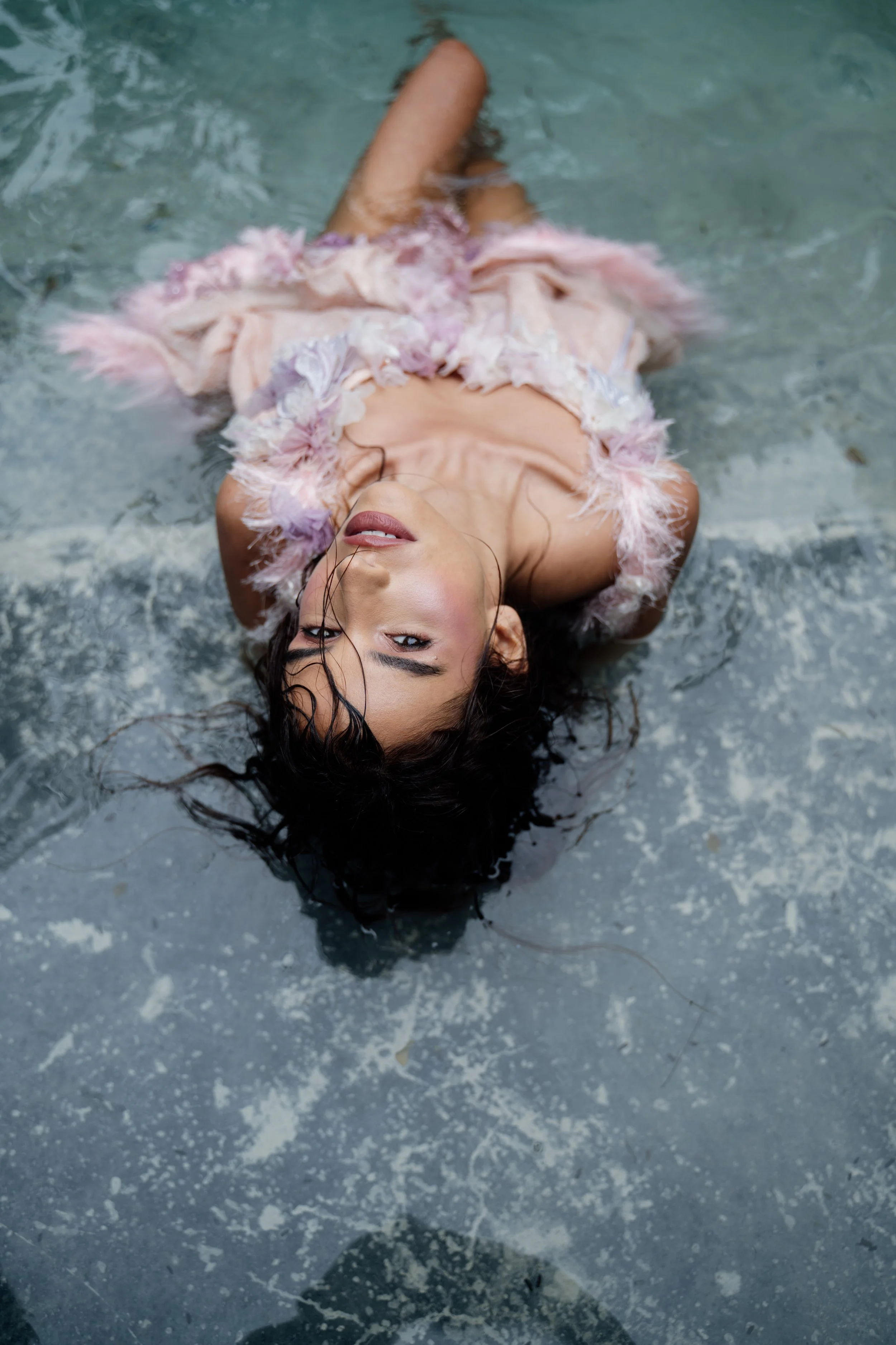 A woman lying on a wet surface, wearing a pink, feather-trimmed outfit, with her head tilted back and eyes closed.