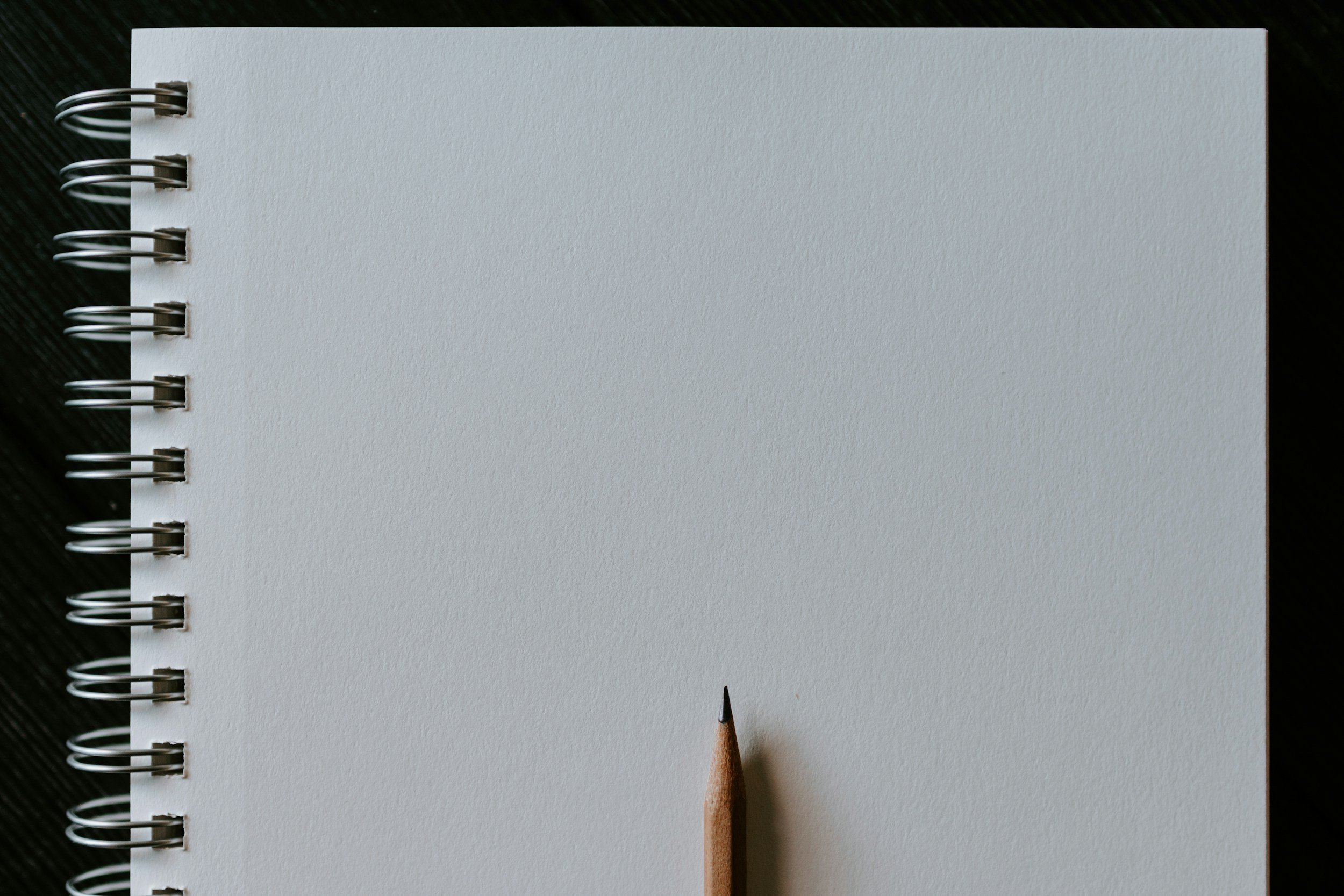 Open spiral notebook with blank white pages and a sharpened pencil at the bottom center on a dark surface.