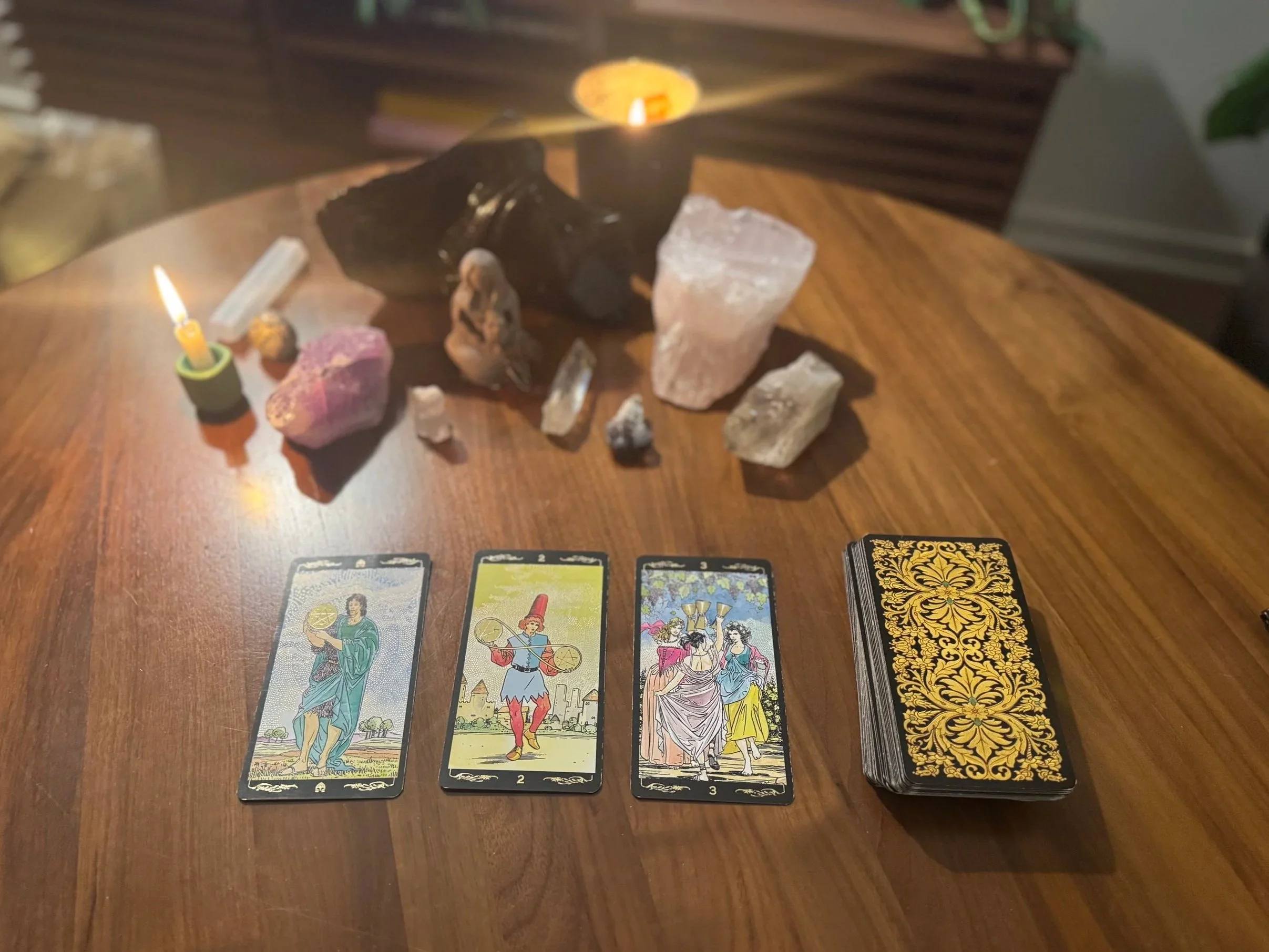 A wooden table with Tarot cards laid out, crystals, candles, and rocks arranged for a reading or spiritual practice.