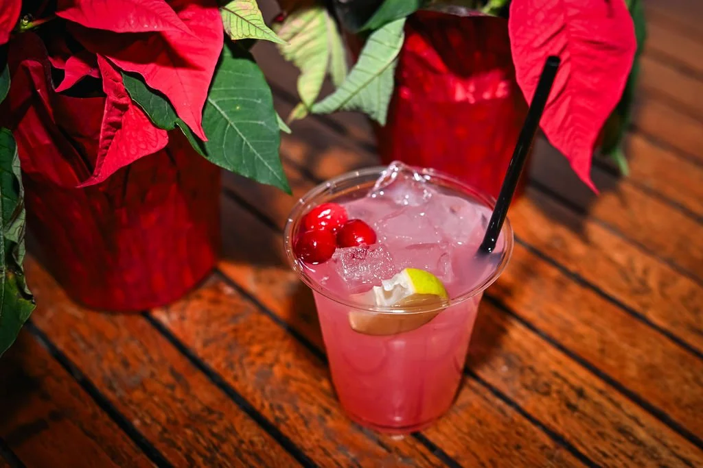 Pink cocktail with ice, cherry garnish, lemon slice, and a black straw on a wooden table, with poinsettia plants in red pots in the background.