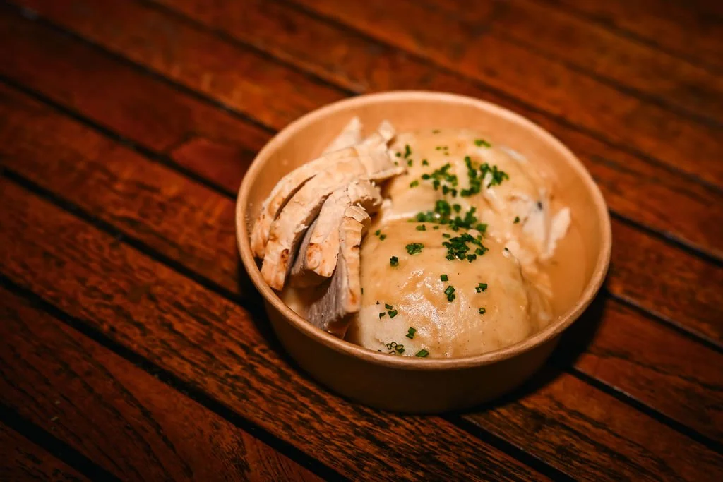 A bowl of chicken and mashed potatoes with gravy, garnished with chopped green herbs, on a wooden surface.
