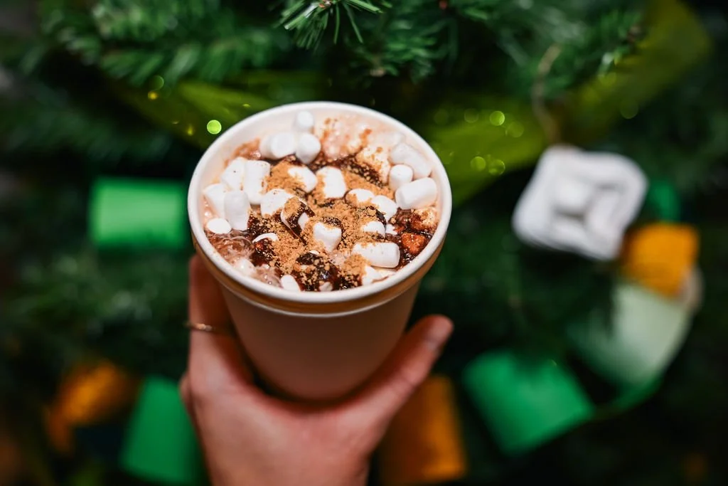 A hand holding a cup of hot chocolate topped with mini marshmallows and cocoa powder, with a decorated Christmas tree in the background.