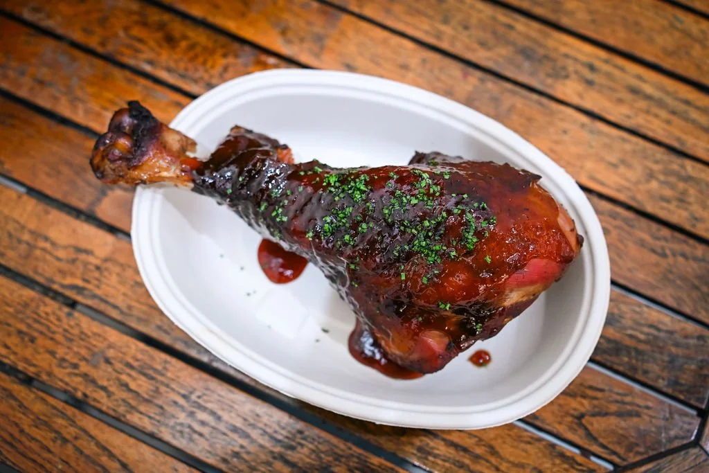 Barbecue pork knuckle with sauce and chopped herbs on a white oval plate, wooden table background.
