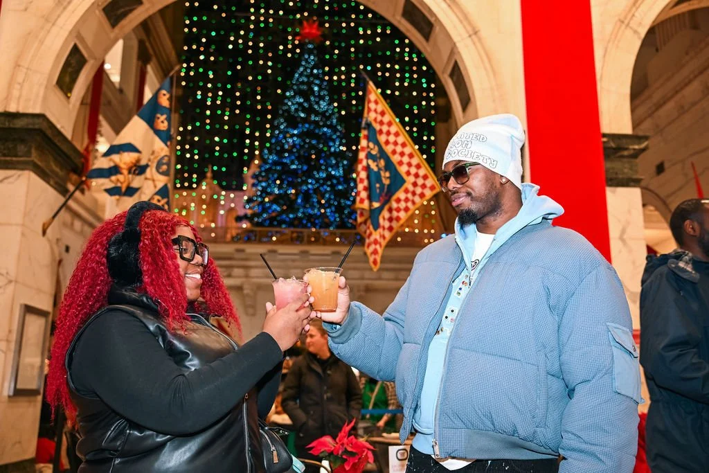 A man and woman toasting with drinks in front of a decorated Christmas tree inside a building with arches and holiday banners.