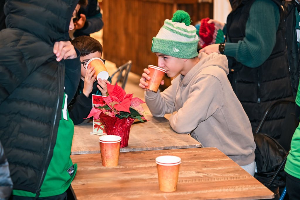 Two children in winter clothing drinking from disposable cups at a table decorated with a poinsettia plant, with other people around.
