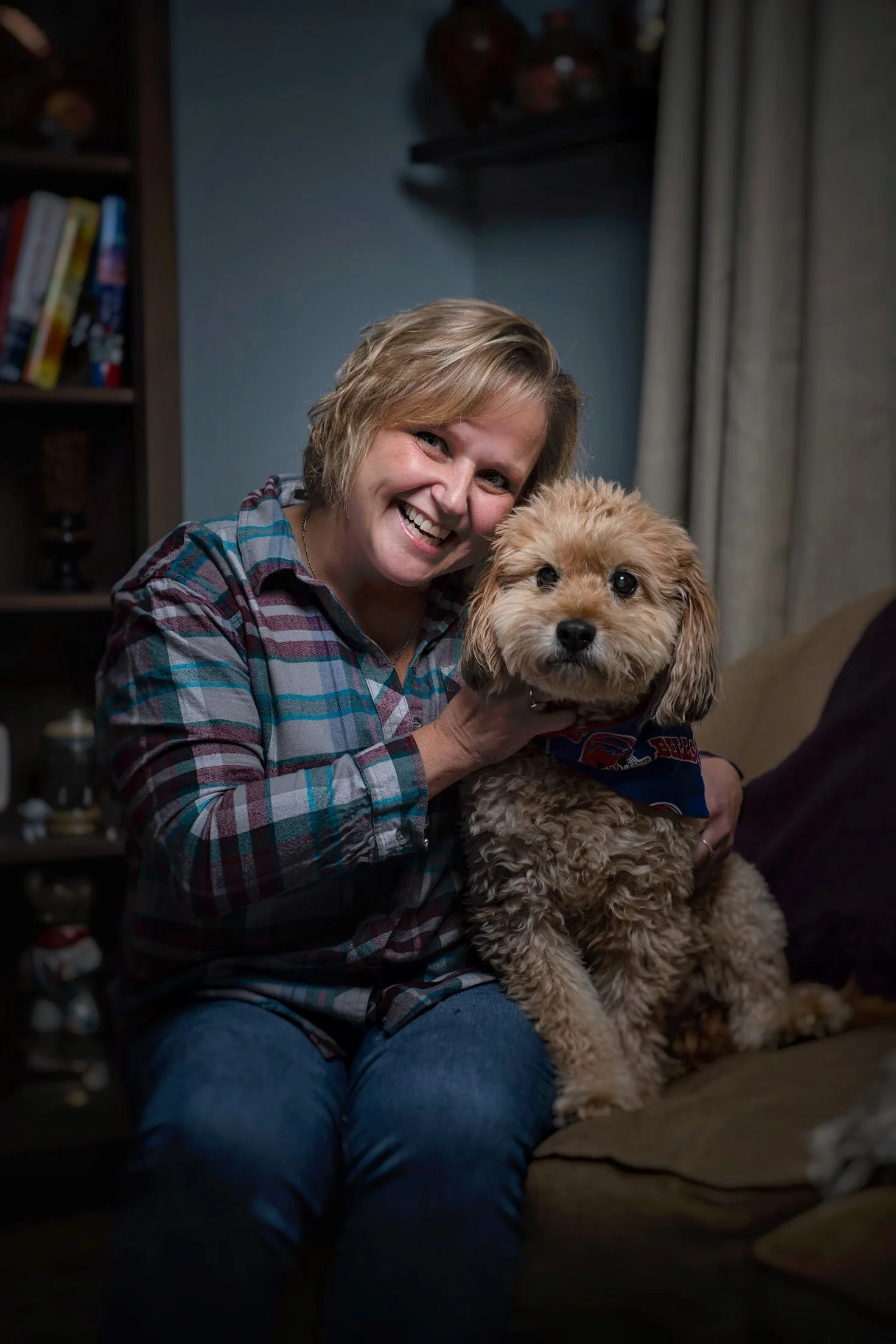 A smiling woman with short blond hair holding a small, fluffy tan dog on a brown couch in a living room.