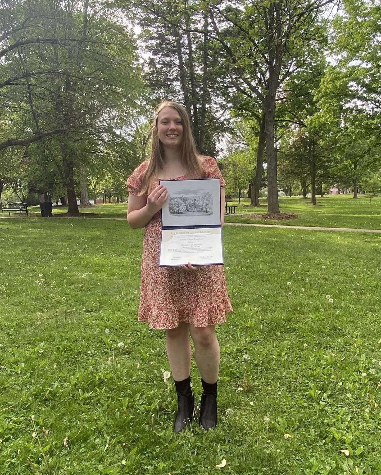 Young woman holding a diploma and a photograph while standing on a grassy park with trees.