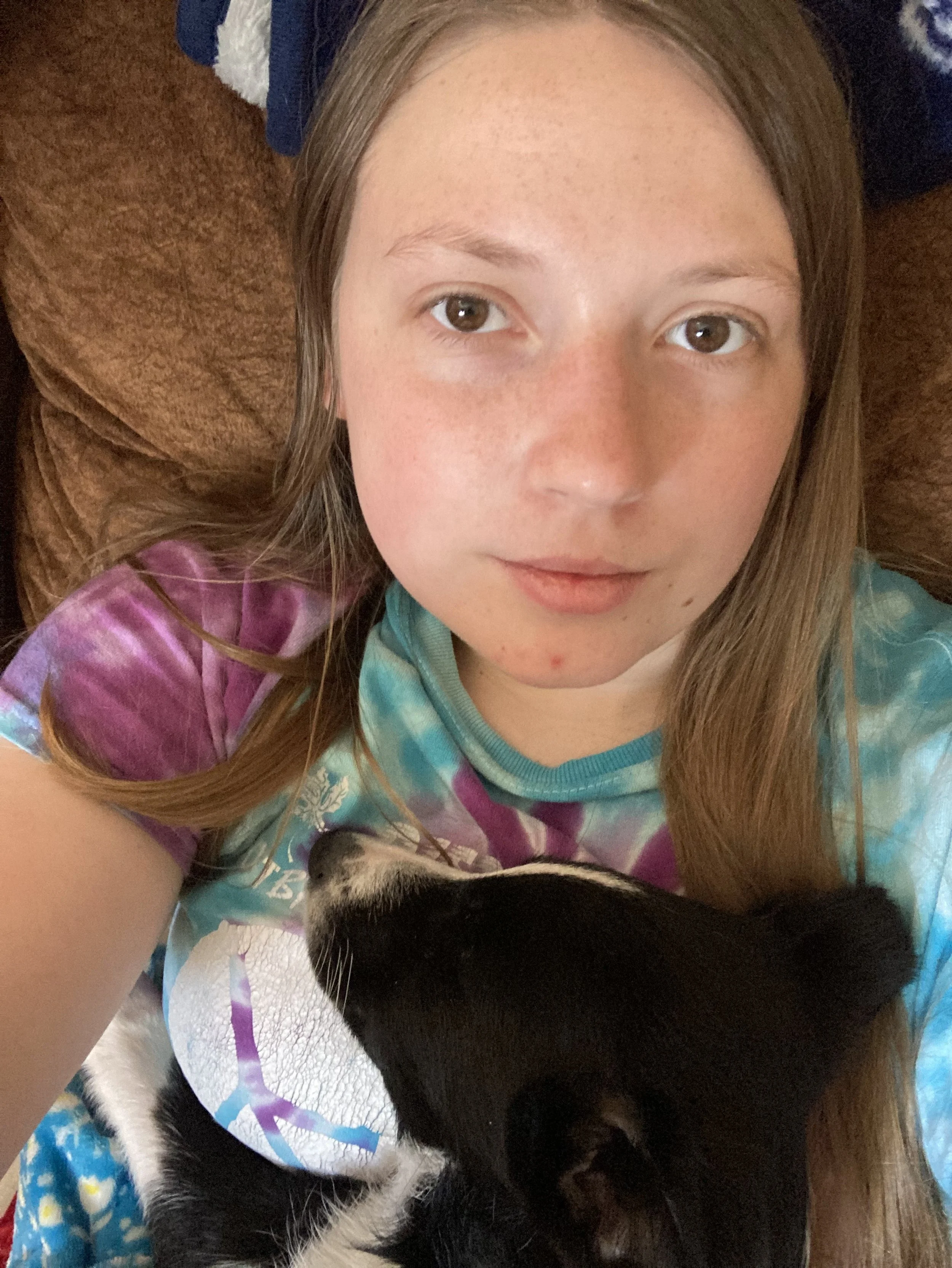 A girl with light skin and long brown hair lying on a brown couch, taking a selfie with a black and white dog who is resting its head on her chest.