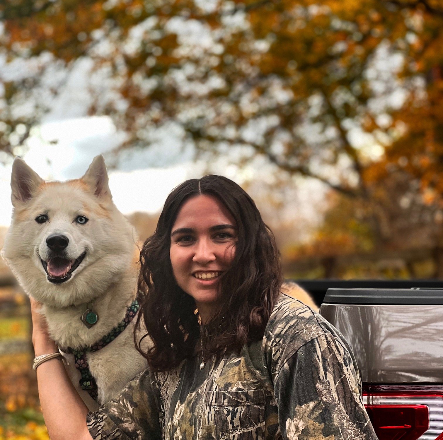 A woman with dark curly hair smiling and holding a white and tan Siberian Husky dog outdoors in a park during fall, with trees with orange and yellow leaves in the background and a black truck nearby.