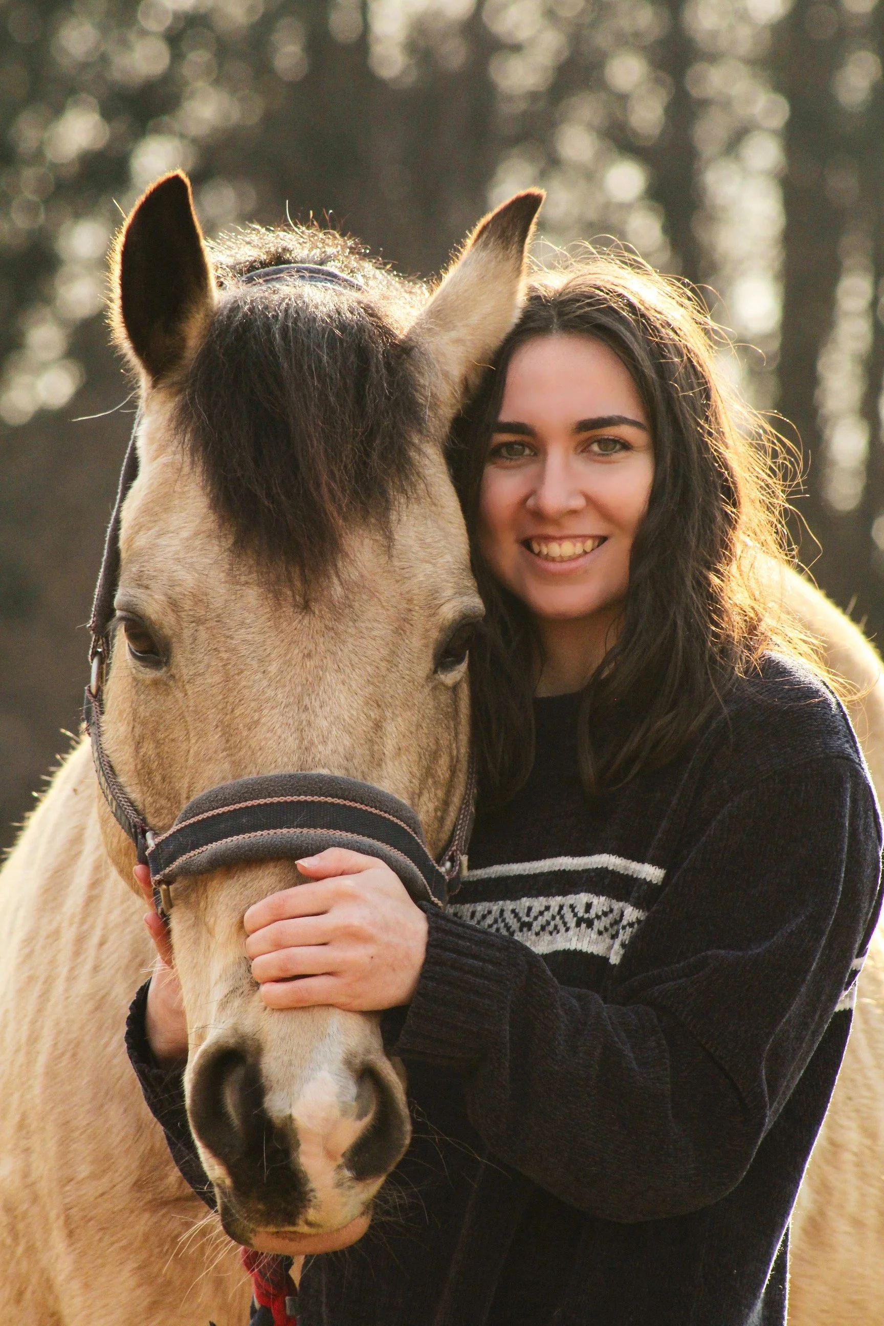 A young woman with long dark hair smiling and hugging a light brown horse with a black mane, outdoors in a wooded area.