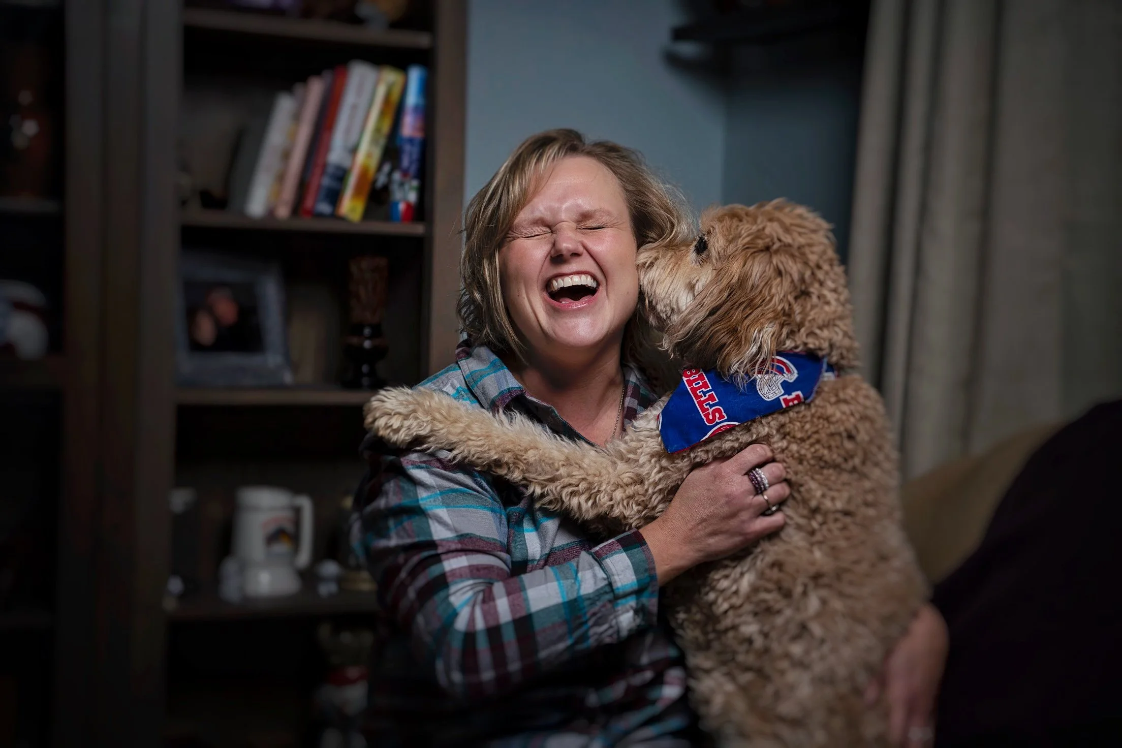 A woman with short hair and a plaid shirt, smiling and laughing, hugging a golden doodle puppy with a blue bandana, in a room with bookshelves and curtains.