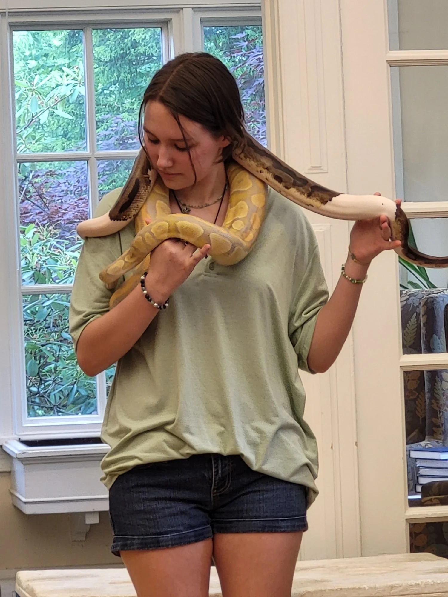 A young woman holding a large snake in an indoor setting with large windows and greenery outside.