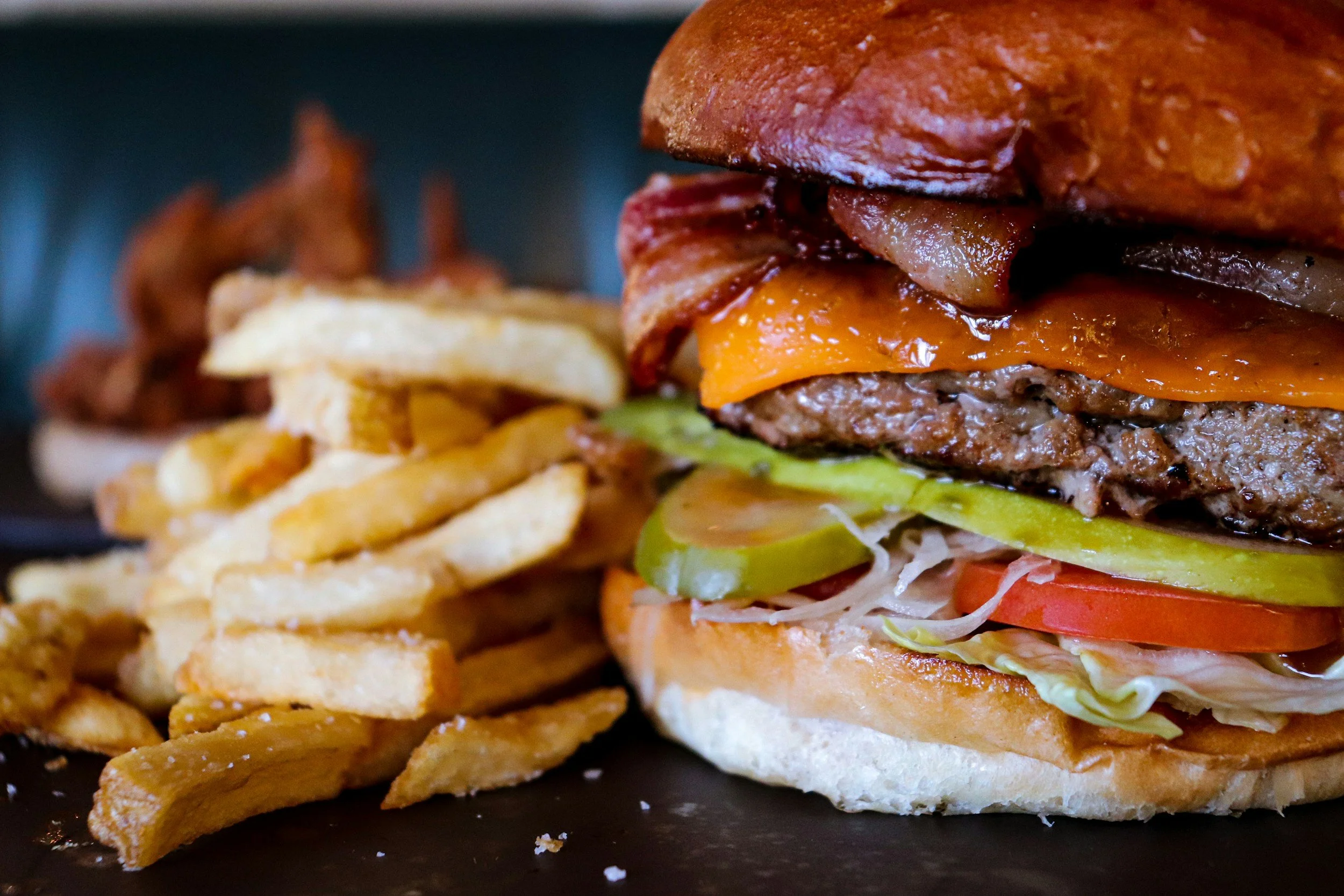 Close-up of a cheeseburger with bacon, lettuce, tomato, pickles, and onions, served with a side of French fries and bacon bits.
