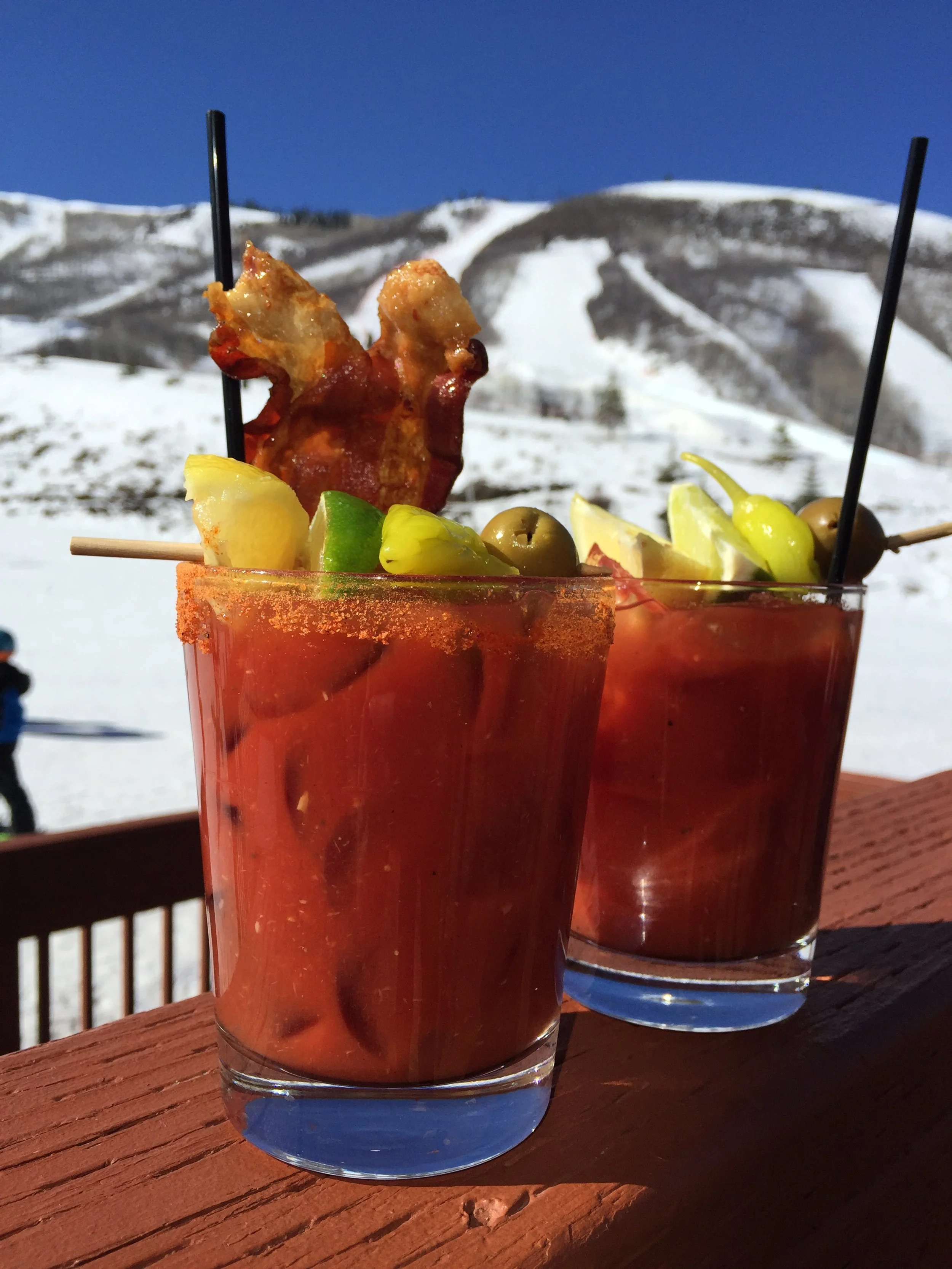 Two glasses of Bloody Mary cocktails with garnishes of skewered bacon, lemon, lime, green olives, and celery, set outdoors with snowy mountain background.