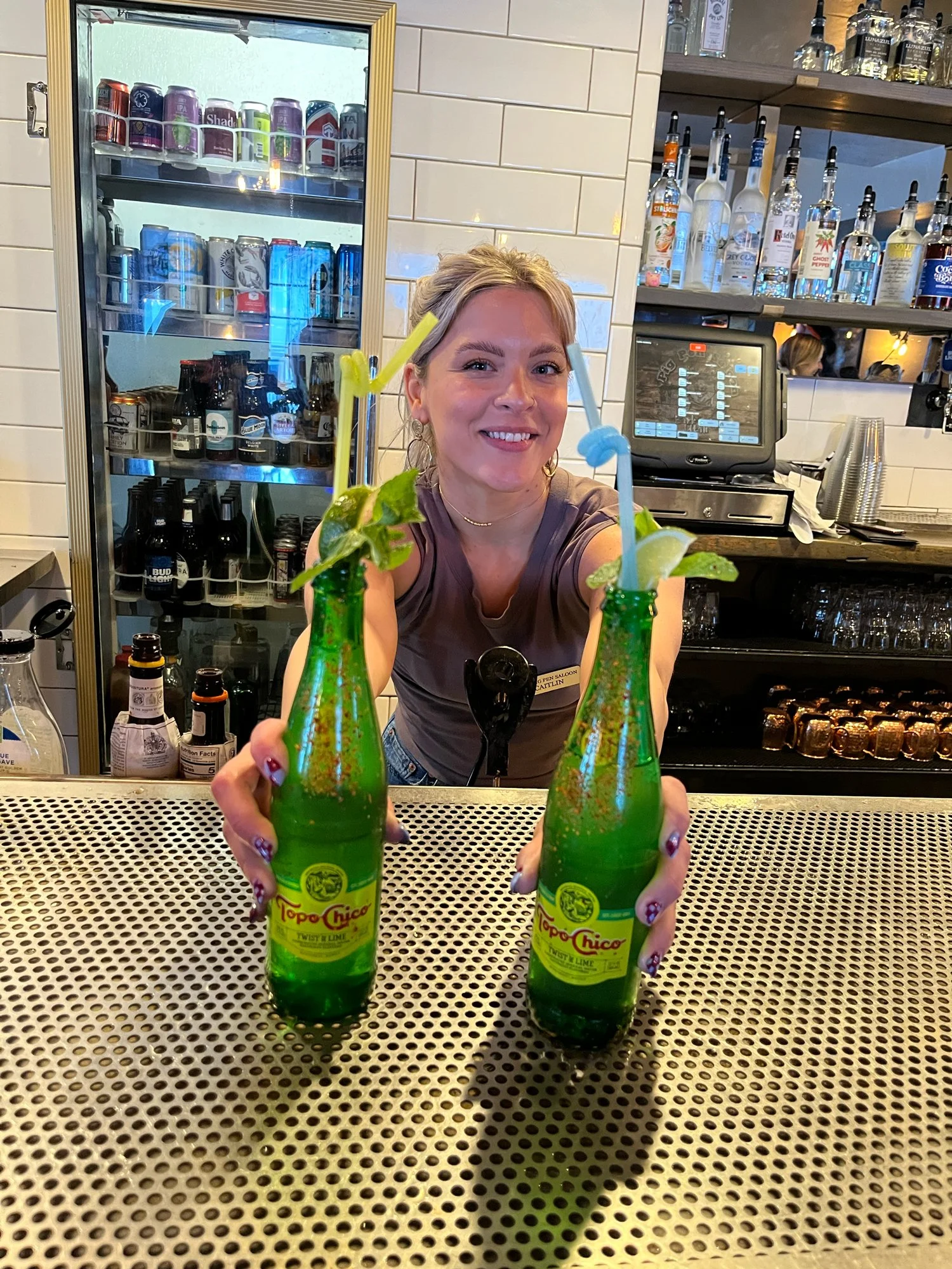 A woman smiling at the camera behind a bar, holding two green bottles of Topo Chico sparkling water, each with a straw and a lime wedge, in a bar or restaurant setting.