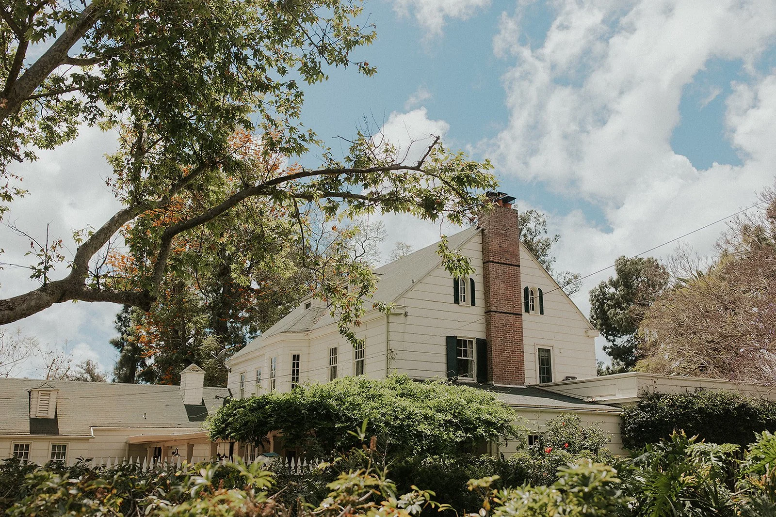 A white, two-story house with a brick chimney, surrounded by lush green shrubs and trees under a partly cloudy sky.
