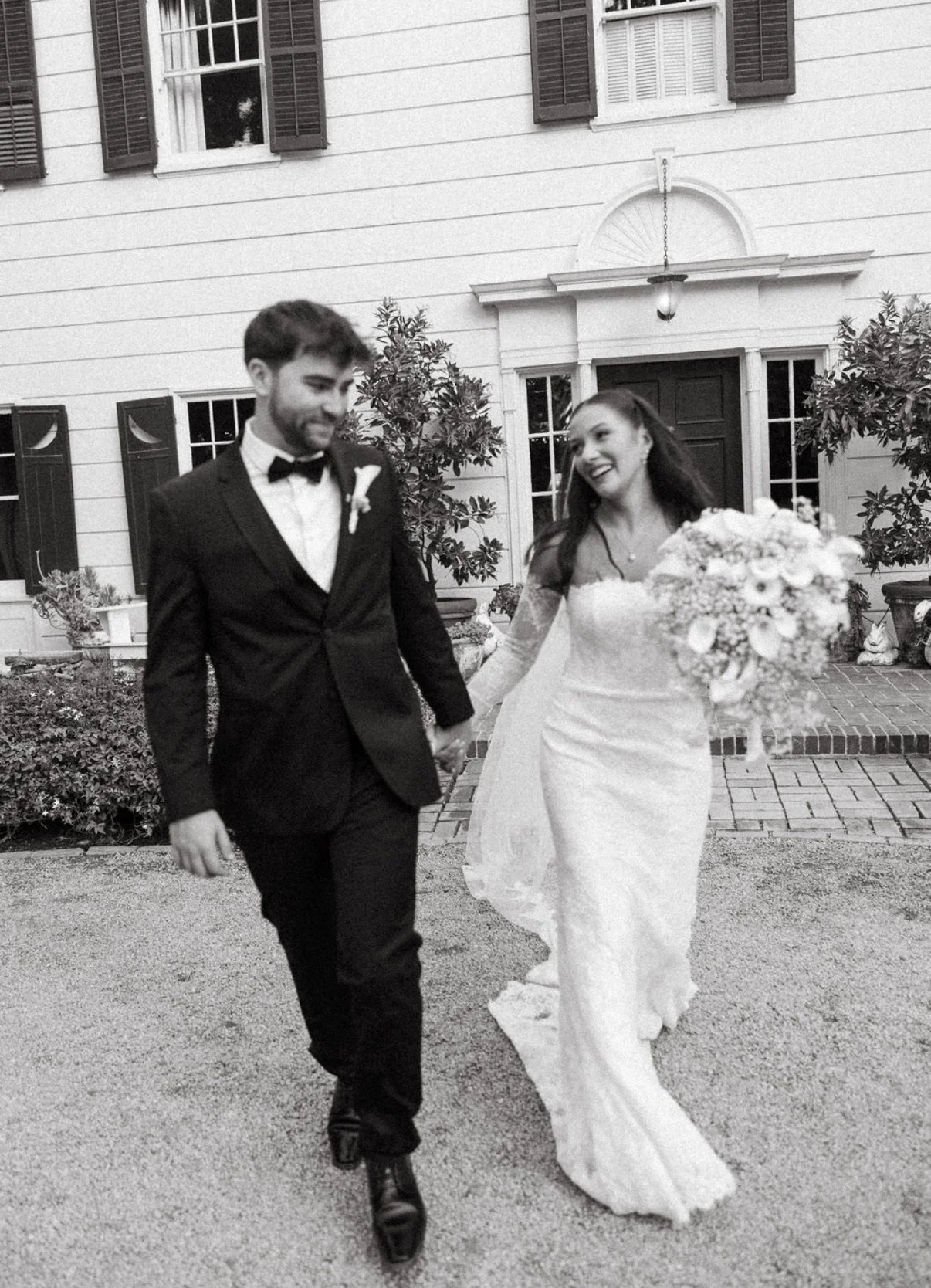 A bride and groom walking hand in hand outside a house, smiling. The bride is wearing a white wedding dress and holding a large bouquet. The groom is wearing a black tuxedo with a bow tie.