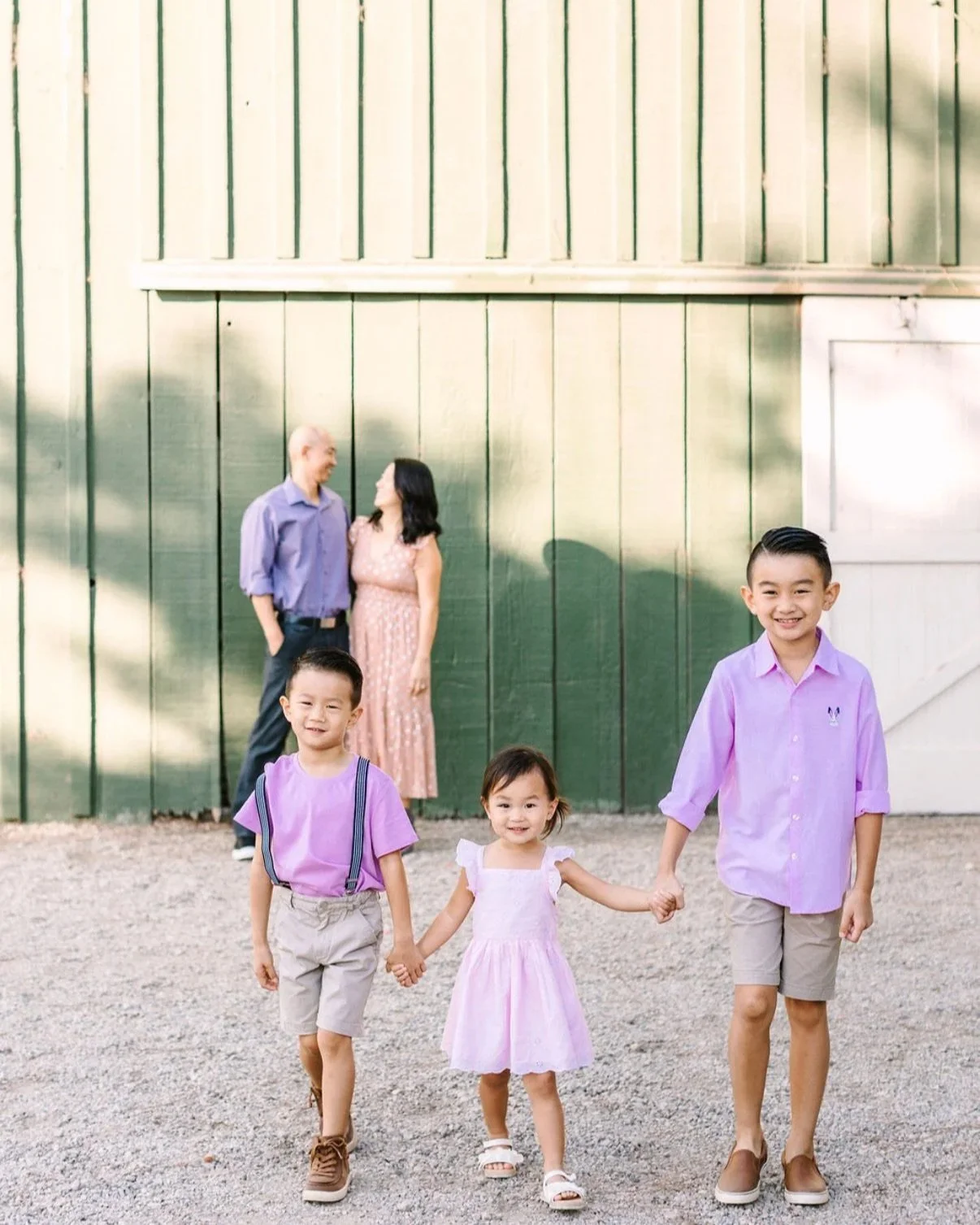 Two boys and one little girl all dressed in matching purple smiling at the camera while their parents share a fond embrace in front of the green barn at McCormick Home Ranch for their annual family photos.