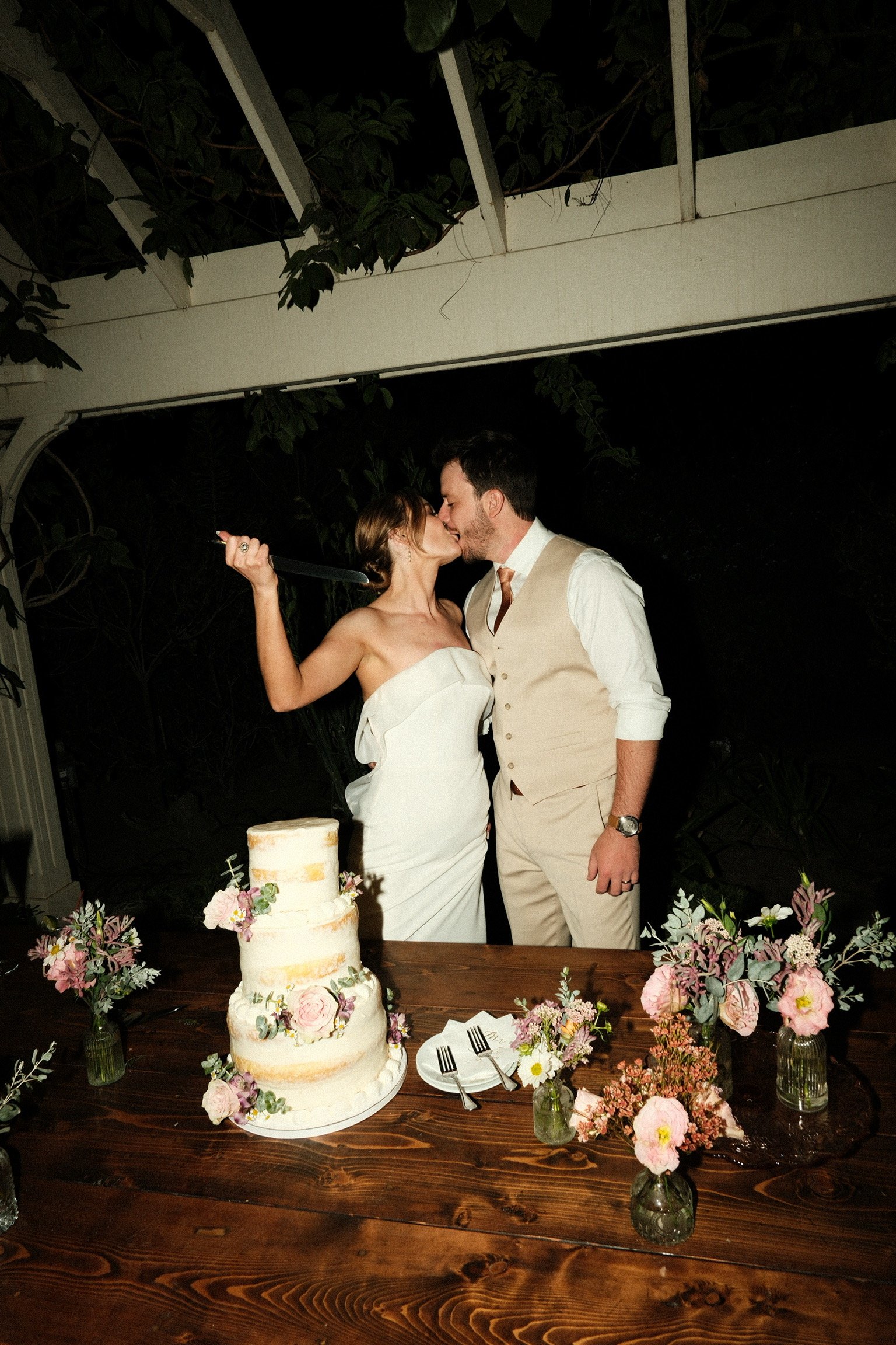 A wedding couple kissing behind a wedding cake decorated with pink flowers, on a wooden table surrounded by small floral arrangements, in an outdoor setting at night.