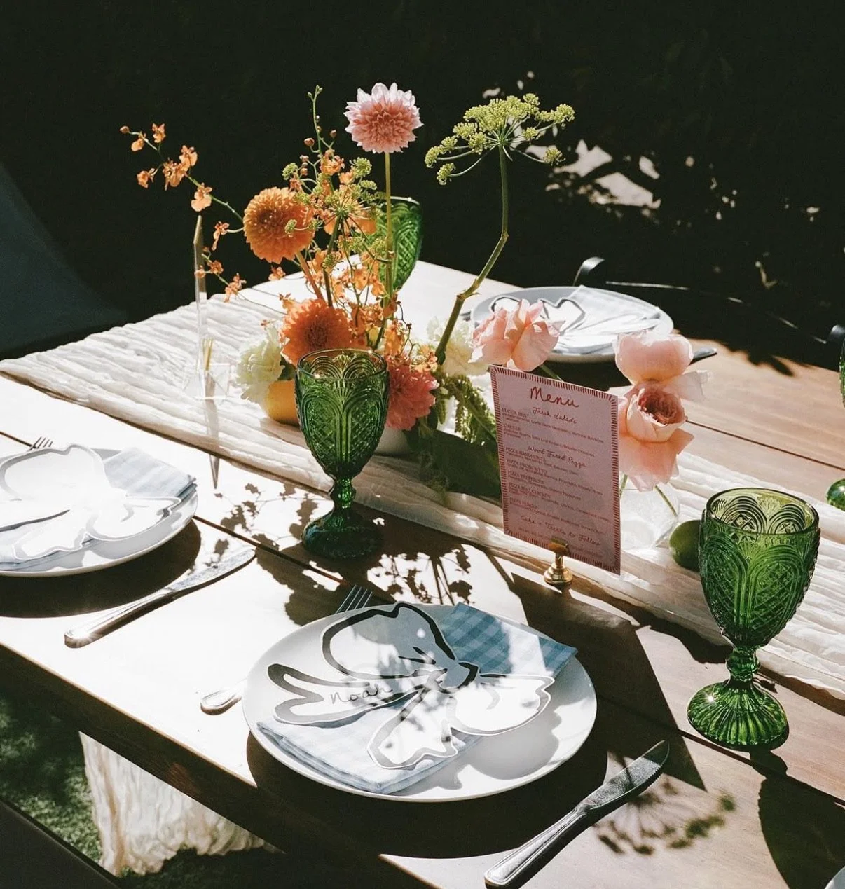 A beautifully set outdoor dining table with white plates, silverware, and green textured glasses. The centerpiece features a floral arrangement with pink, orange, and white flowers in a white vase, along with a pink menu card. Shadow patterns are cas