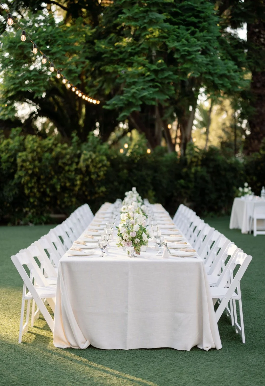 An elegantly decorated outdoor dining table with white chairs, floral centerpiece, glassware, and plates, set in a lush garden with string lights overhead.